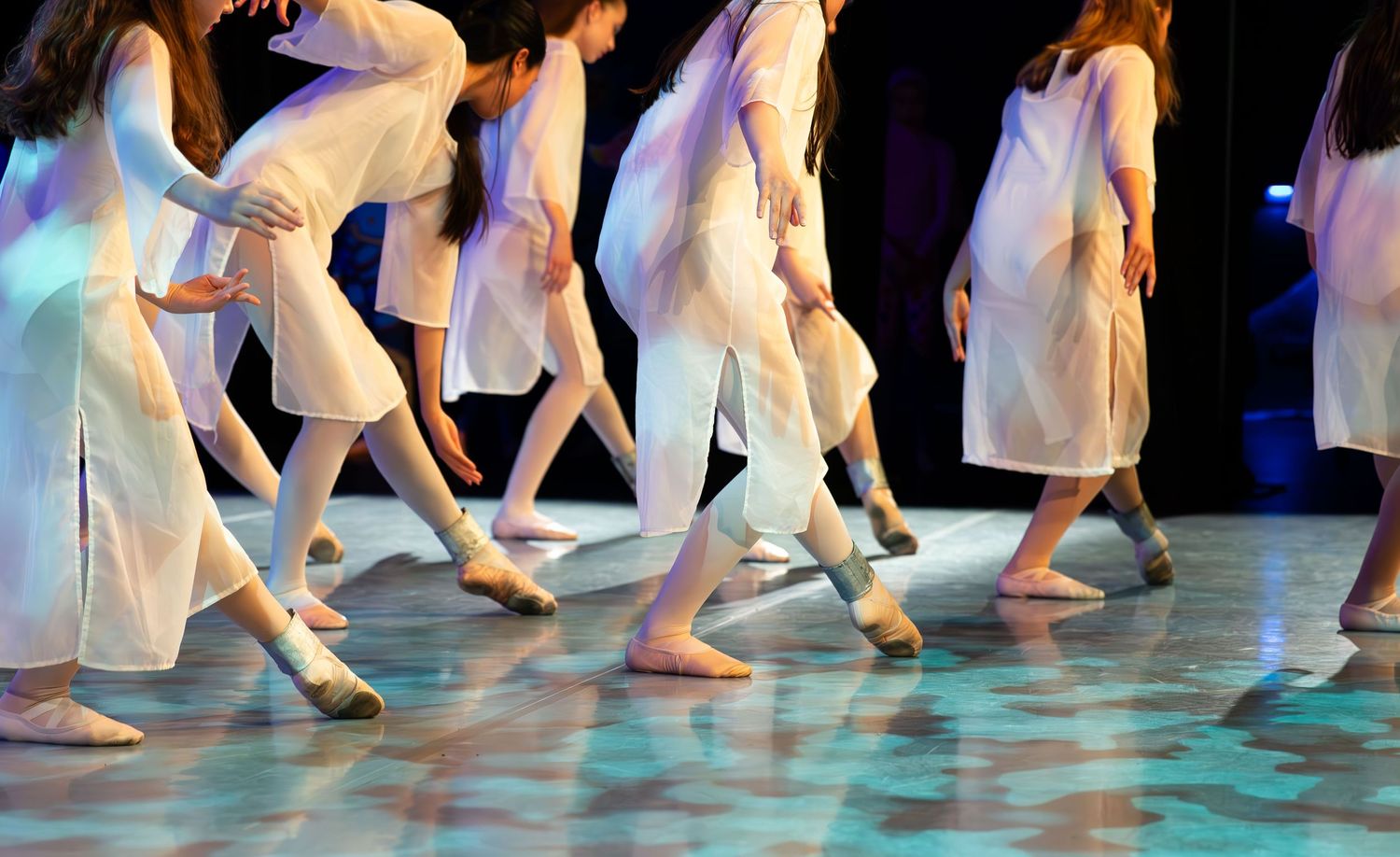 Group of dancers in white costumes performing on a colorful stage, captured mid-movement from the waist down.