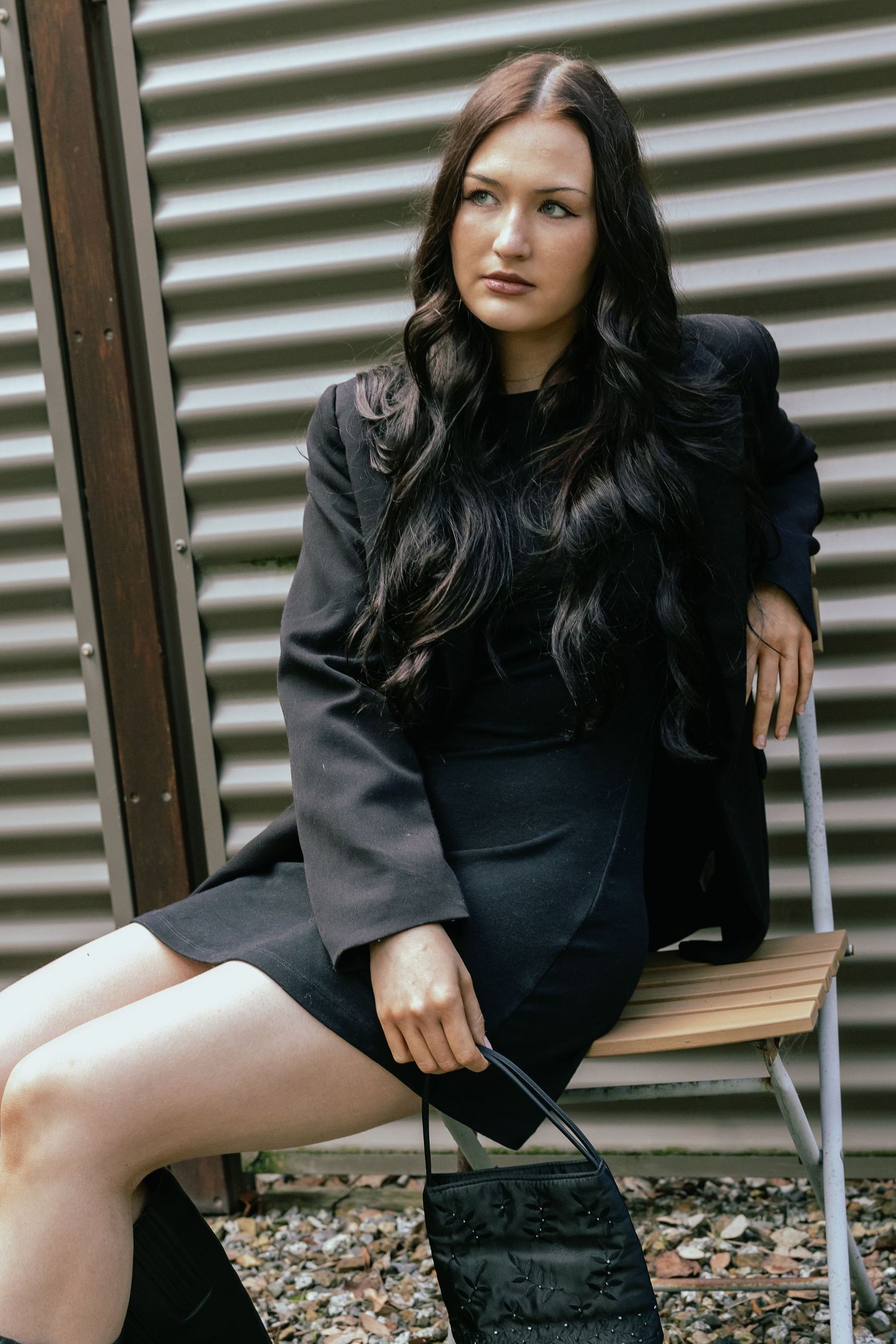 Woman in black blazer and skirt sitting on bench outdoors, holding handbag, with long dark wavy hair.