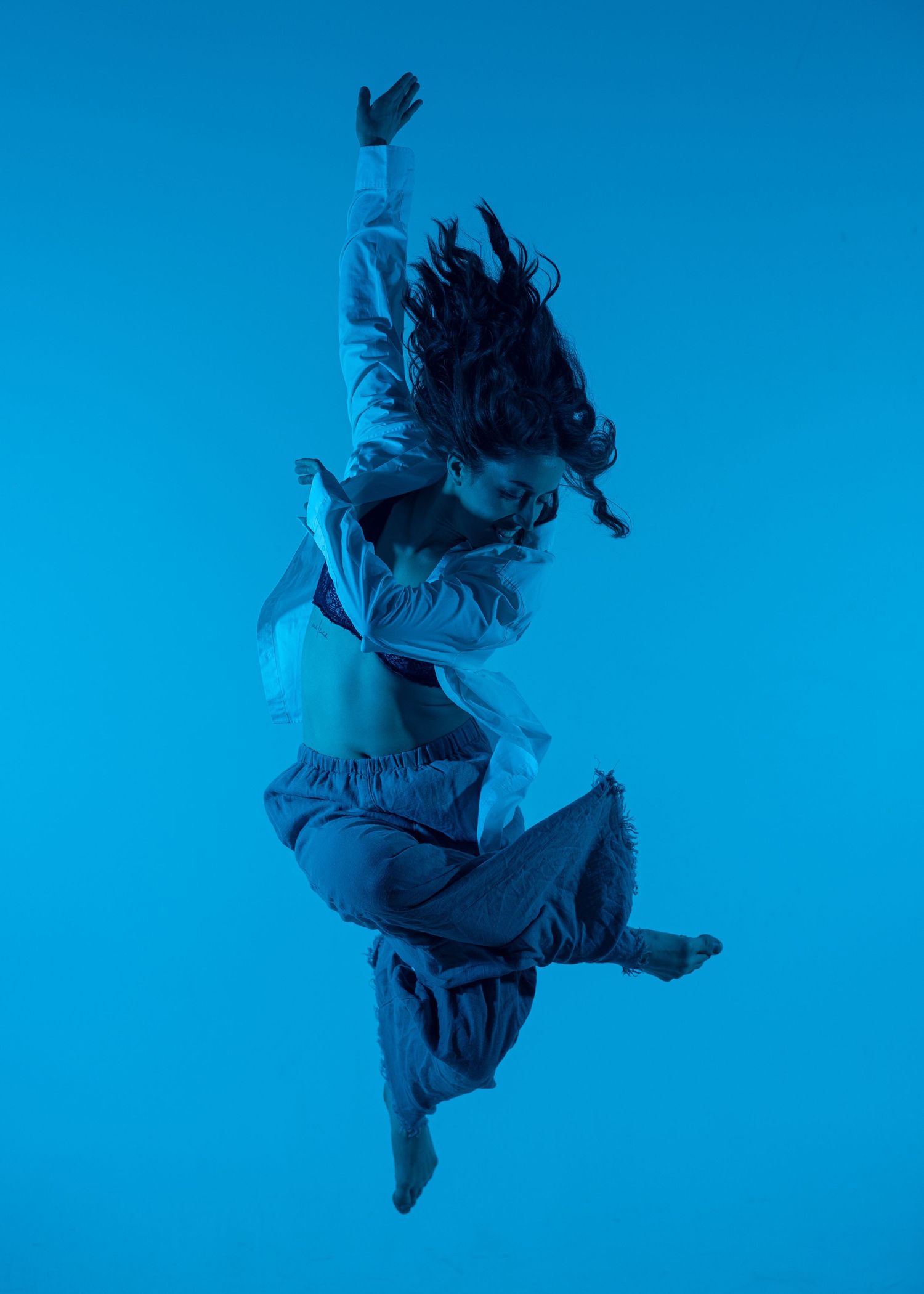 Dynamic female dancer jumping mid-air against a bold blue background in casual white outfit with flowing hair.