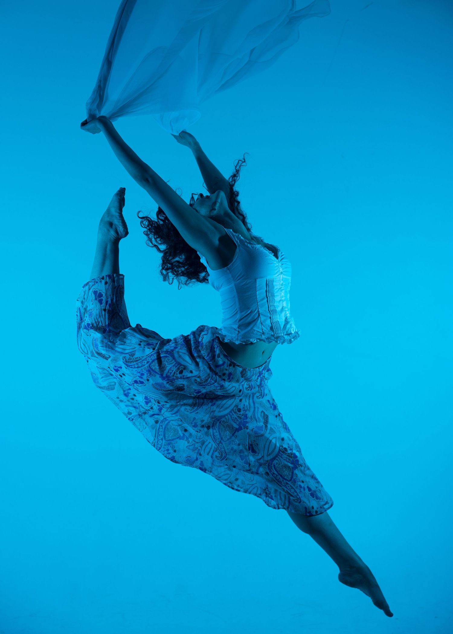 Dancer leaps mid-air against a blue background in a floral skirt, holding flowing white fabric.