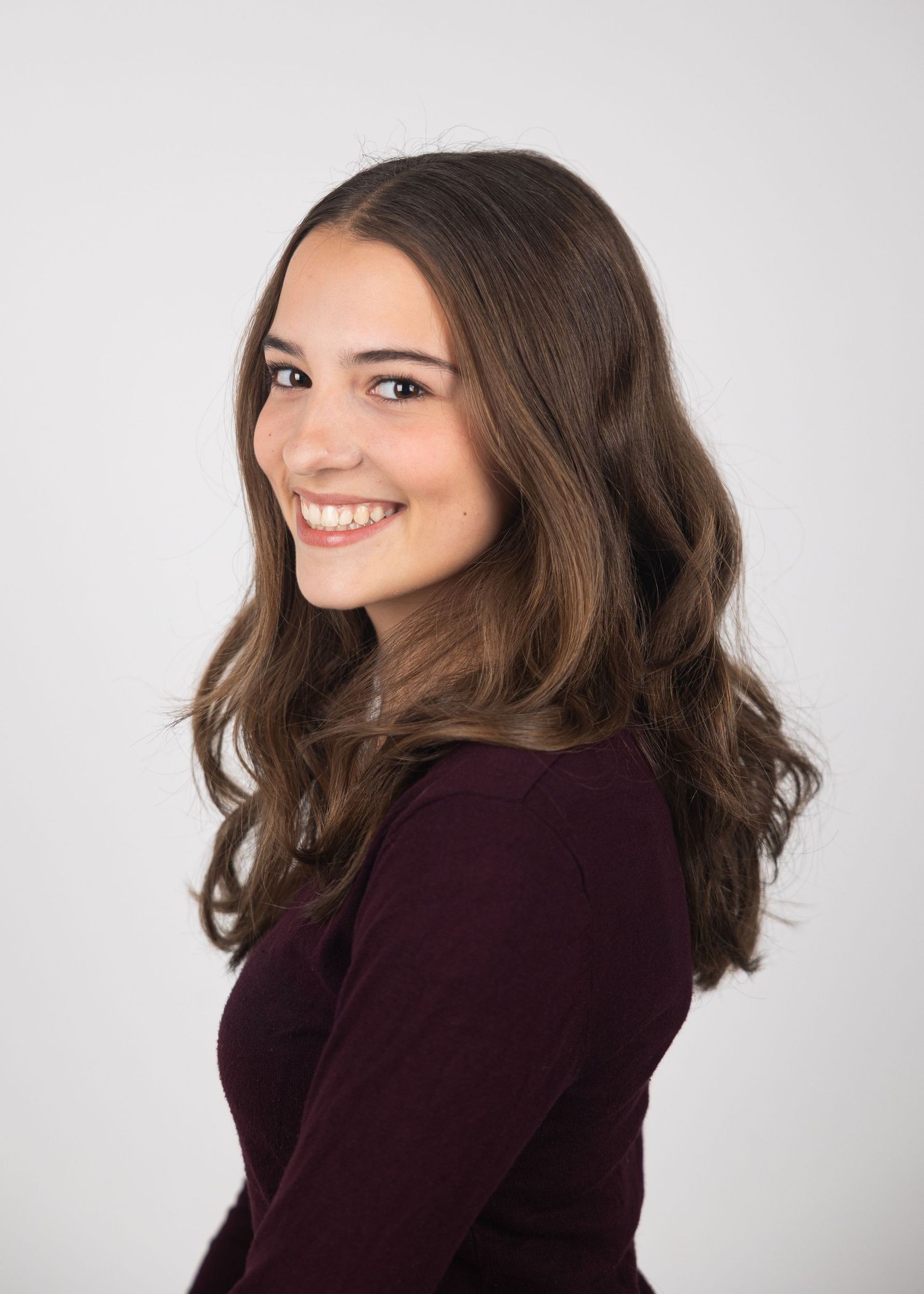 Young woman with long brown wavy hair smiling over shoulder, wearing burgundy top, professional headshot on gray background.