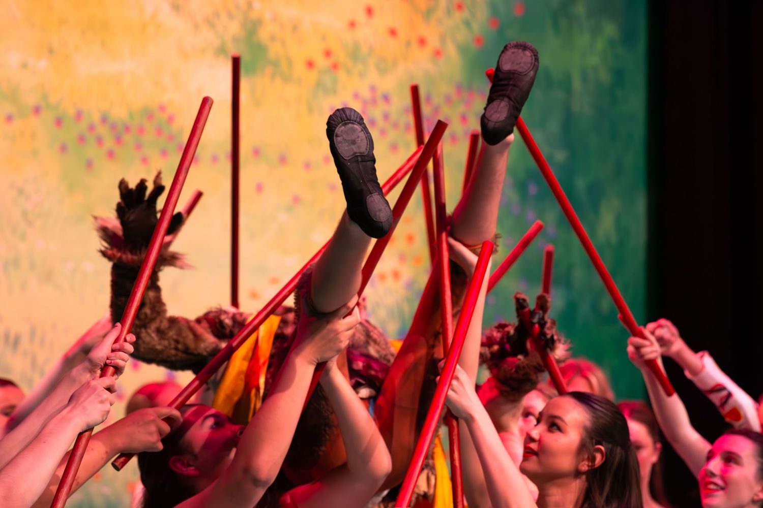 Performers raise red batons and legs in a vibrant choreographed dance routine on a colorful stage.
