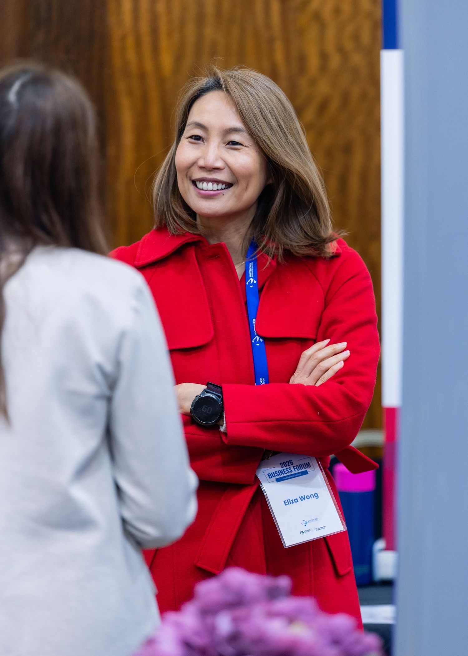 Woman in red jacket with arms crossed smiling at a conference event wearing a name badge.