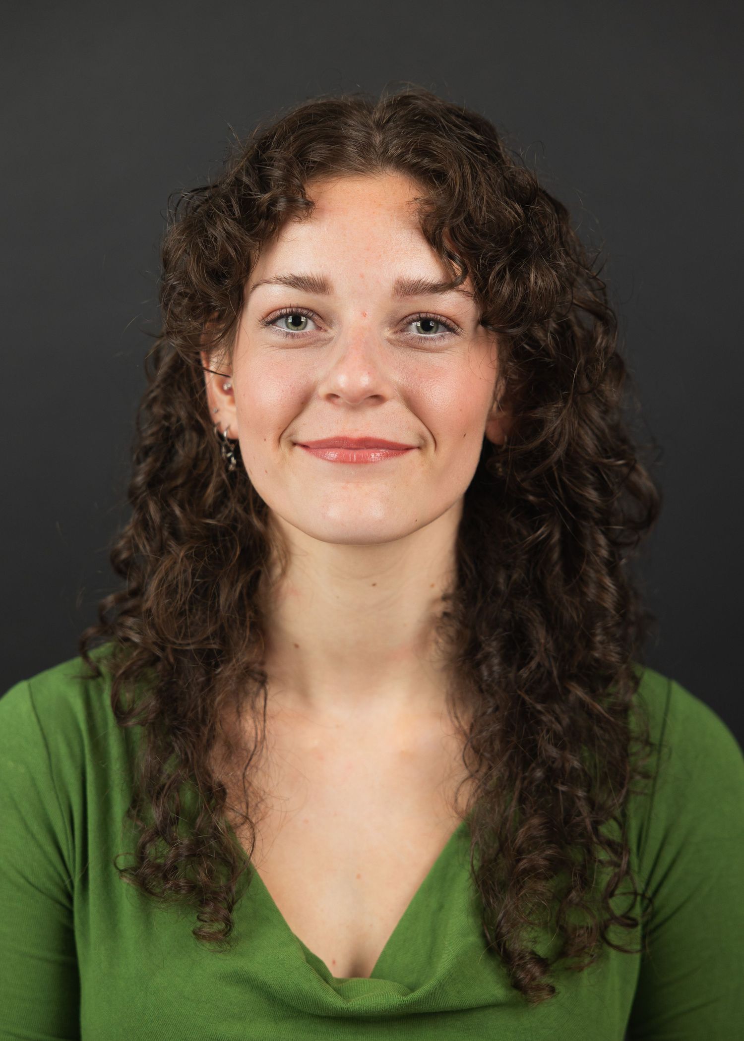 Young woman with curly brown hair smiling warmly, wearing a green top against a dark background.