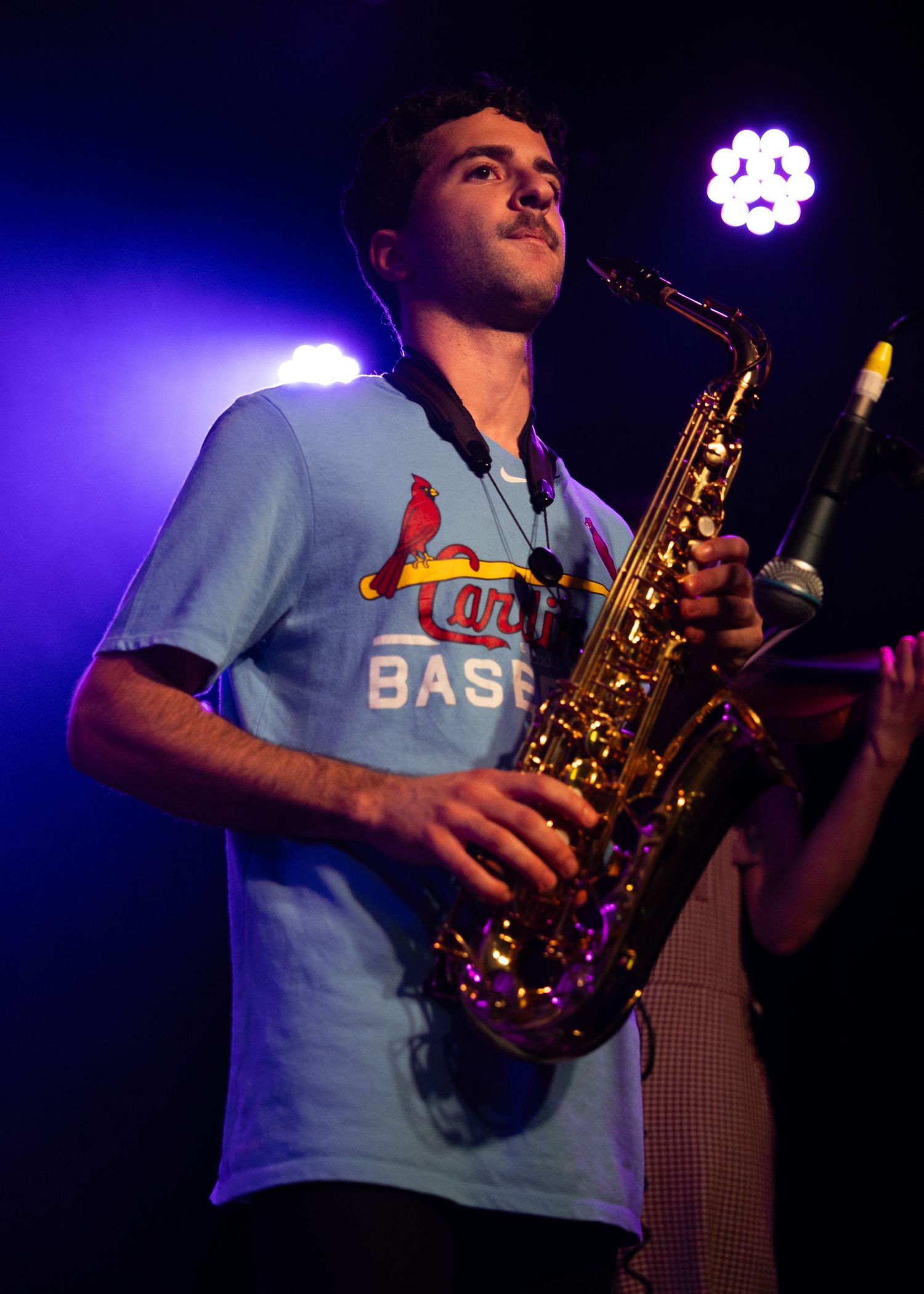 Young man playing alto saxophone on stage wearing a St. Louis Cardinals baseball t-shirt under purple stage lights.