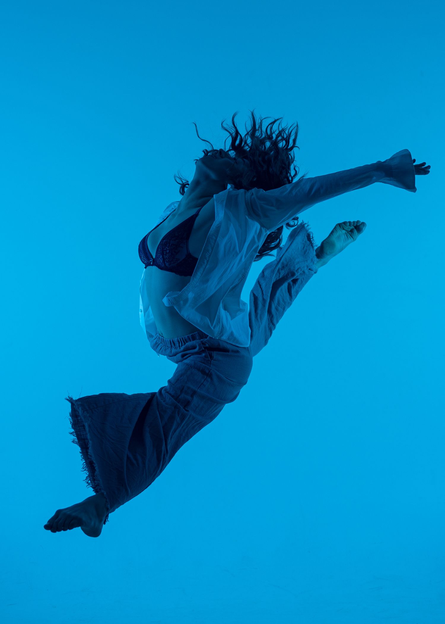 A dancer leaps dynamically against a blue background, wearing wide-leg pants and a white shirt, hair flowing mid-air.