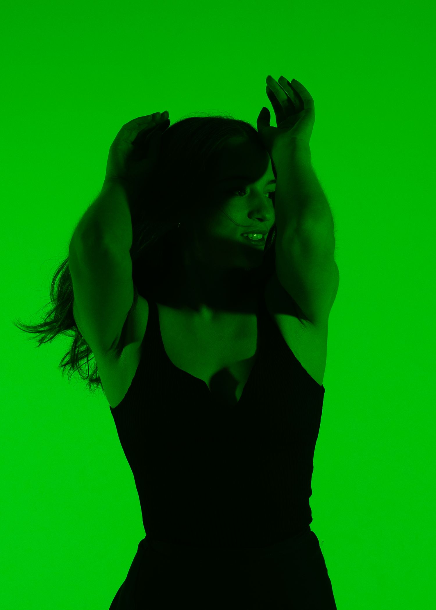 Person dancing against a vibrant green background, arms raised overhead, wearing a black outfit under dramatic green lighting.