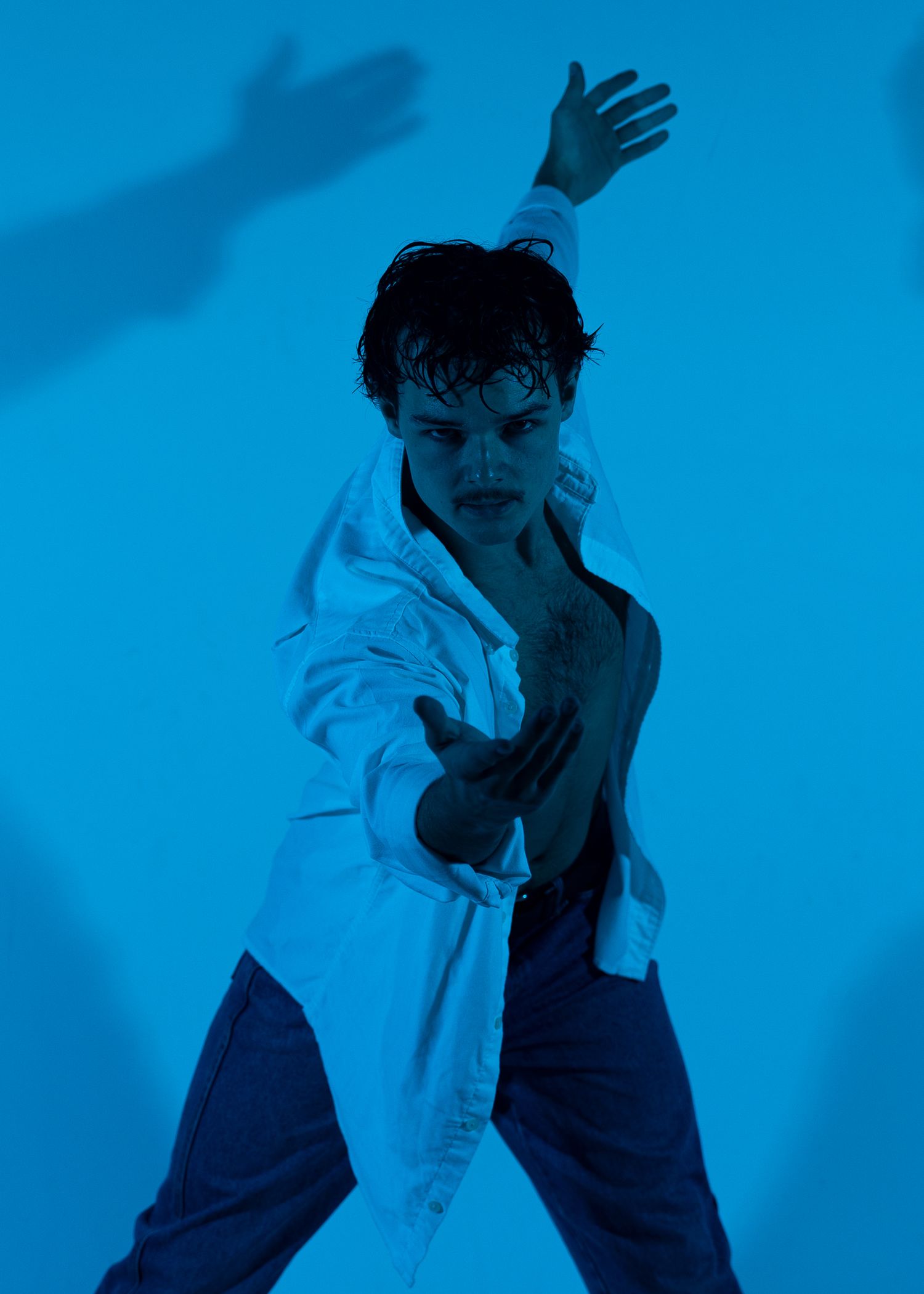 A young man in a white shirt poses dynamically against a blue background with dramatic shadows.