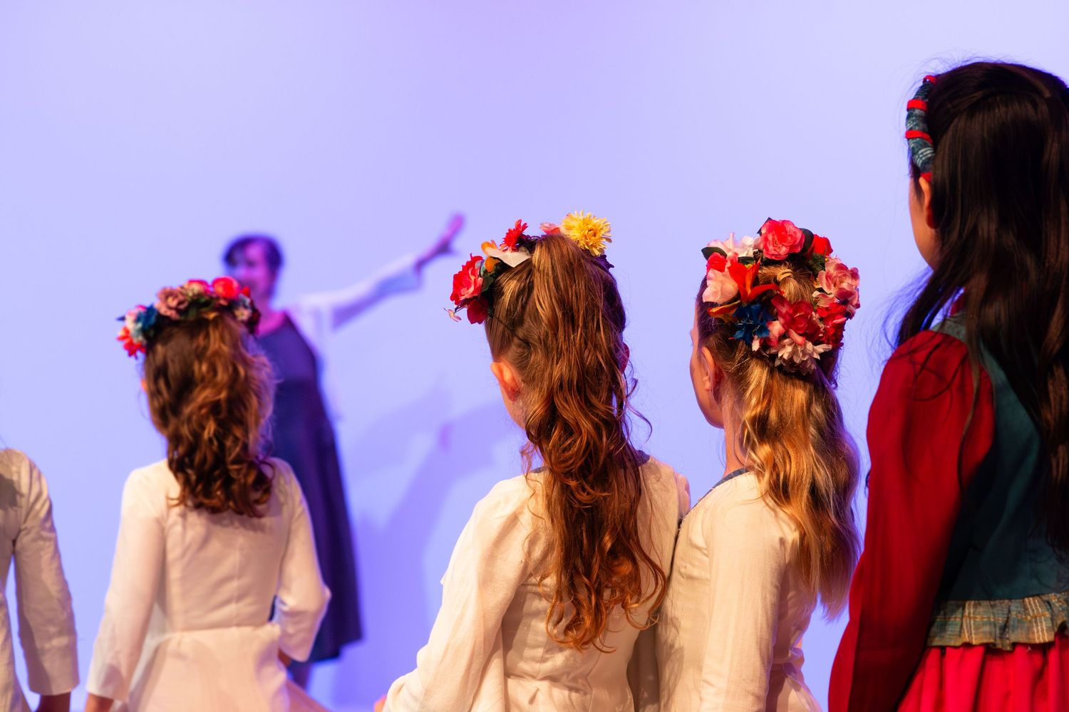 Performers in floral headpieces and long dresses stand in a line on stage with purple lighting behind them.