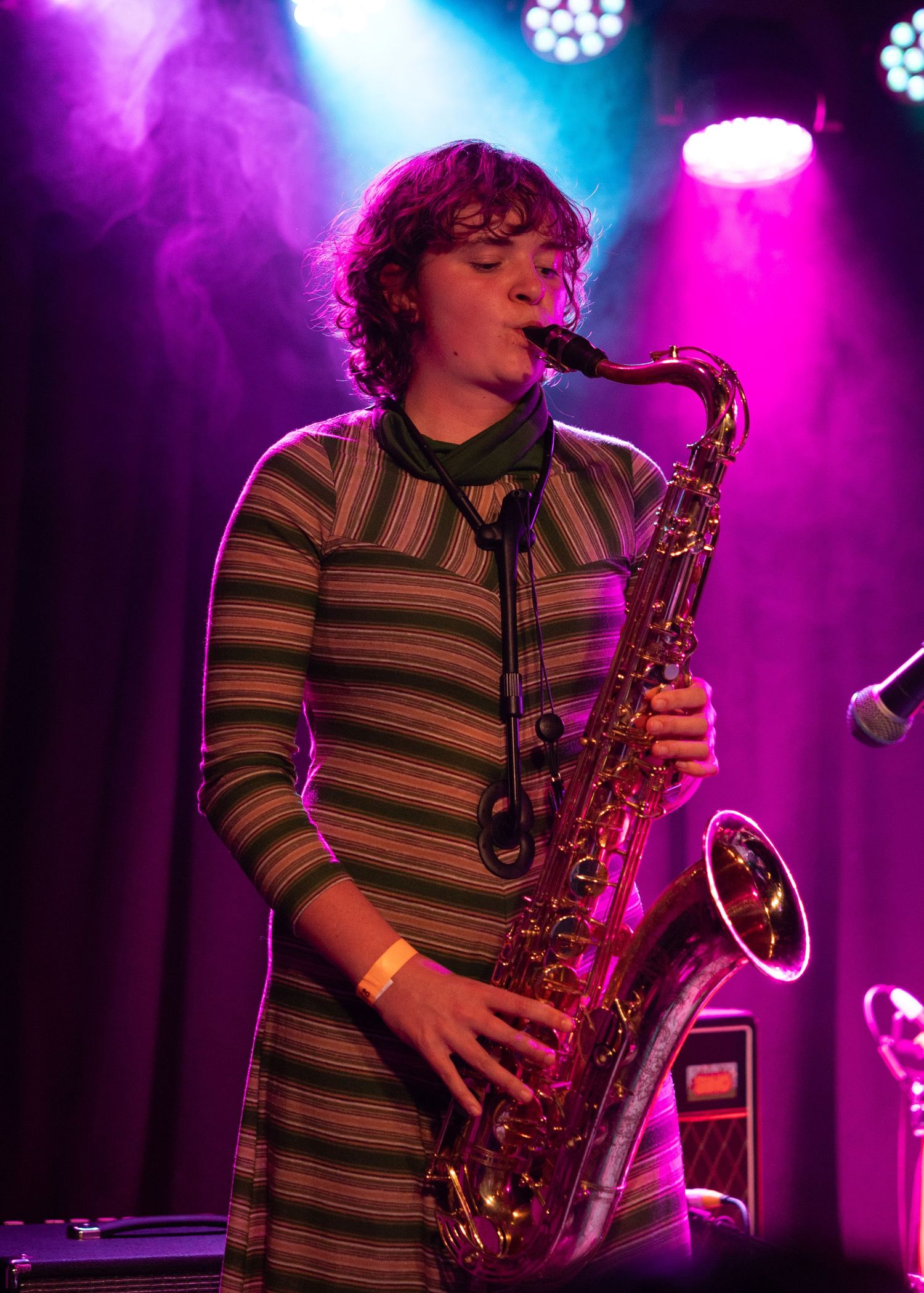 Young woman in striped dress playing saxophone on stage under colorful purple and pink stage lights.