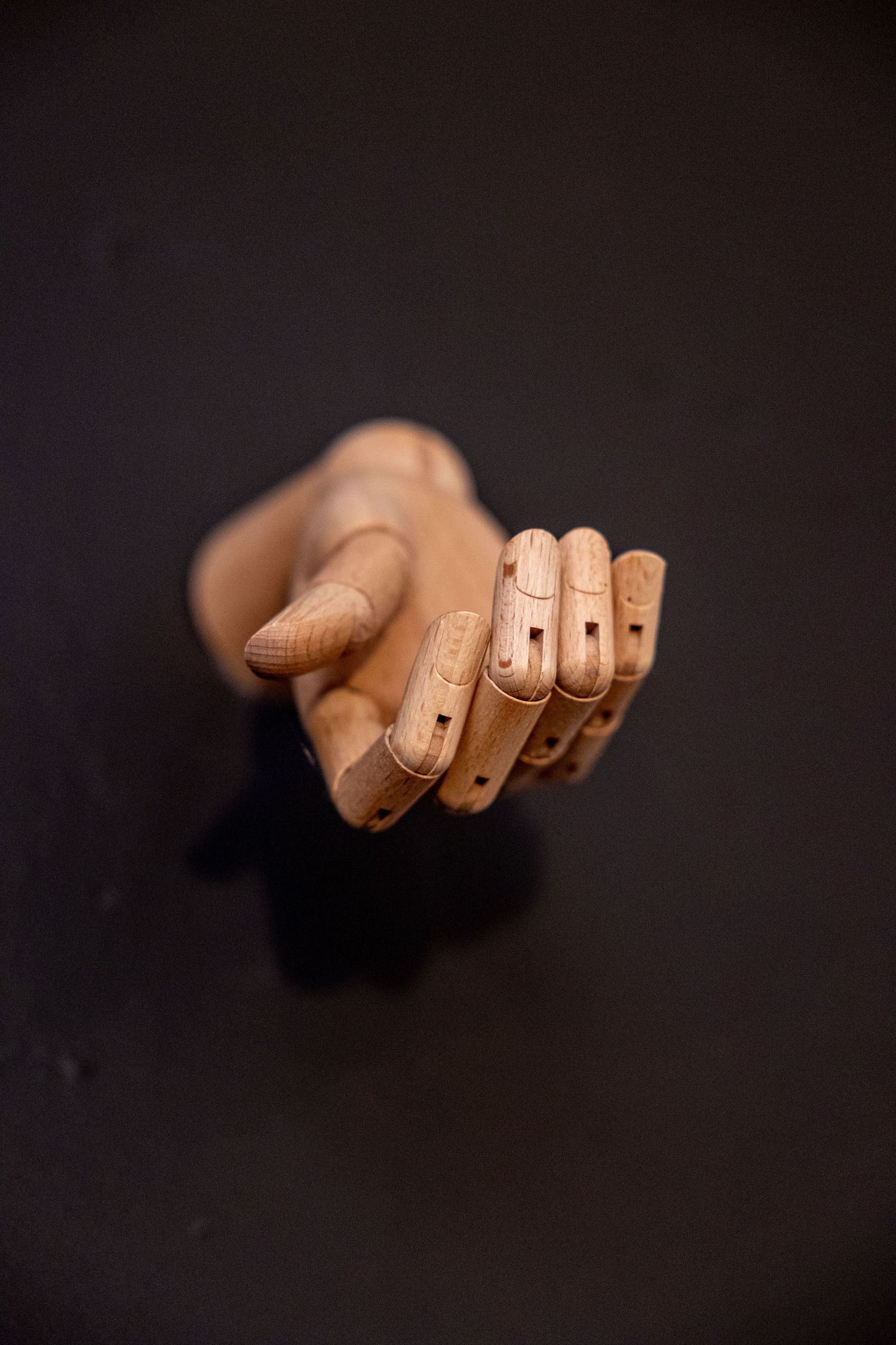 A wooden articulated hand model with jointed fingers pointing downward against a dark background.