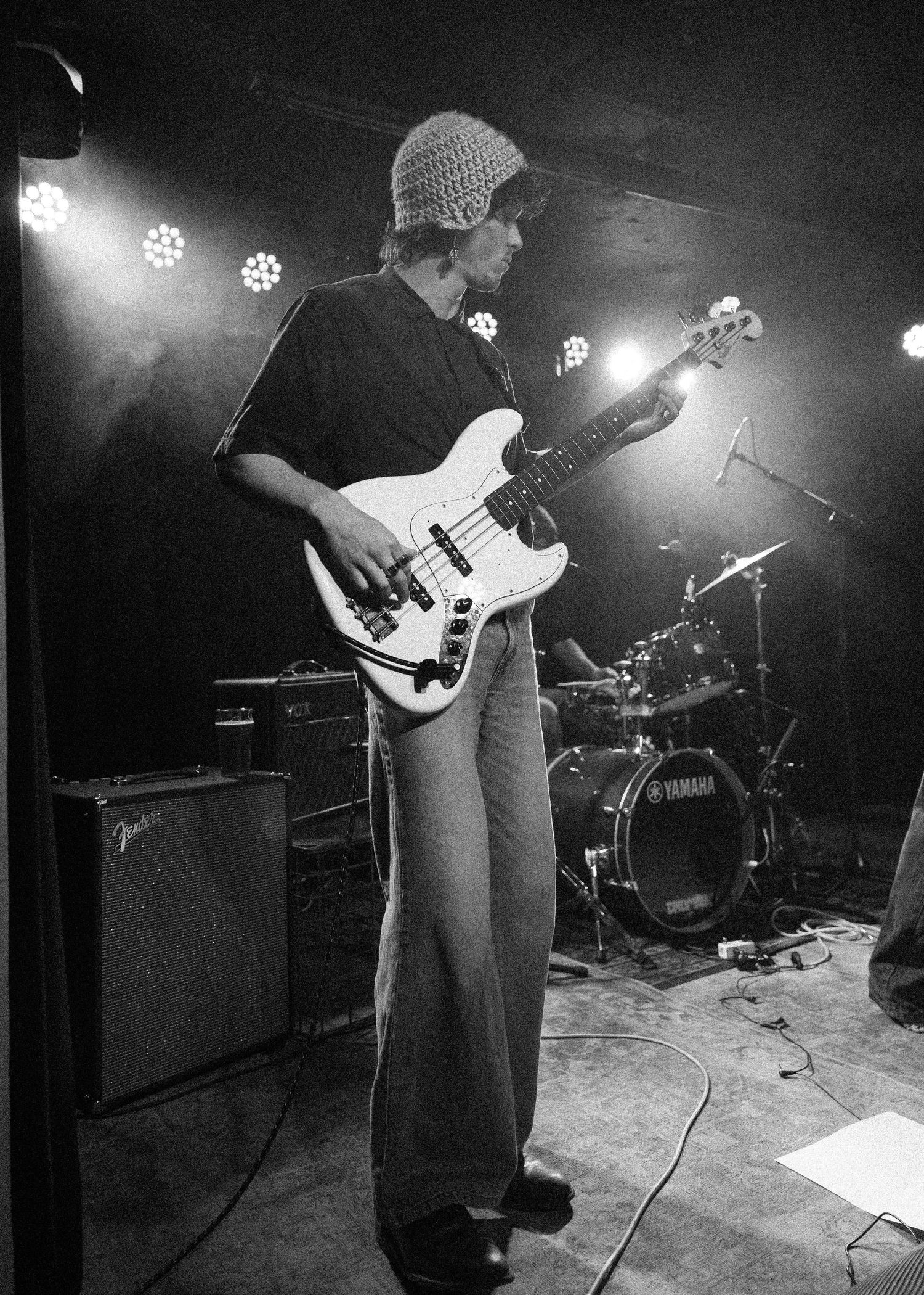 Black and white photo of a musician playing bass guitar on stage with a Yamaha drum kit in the background.