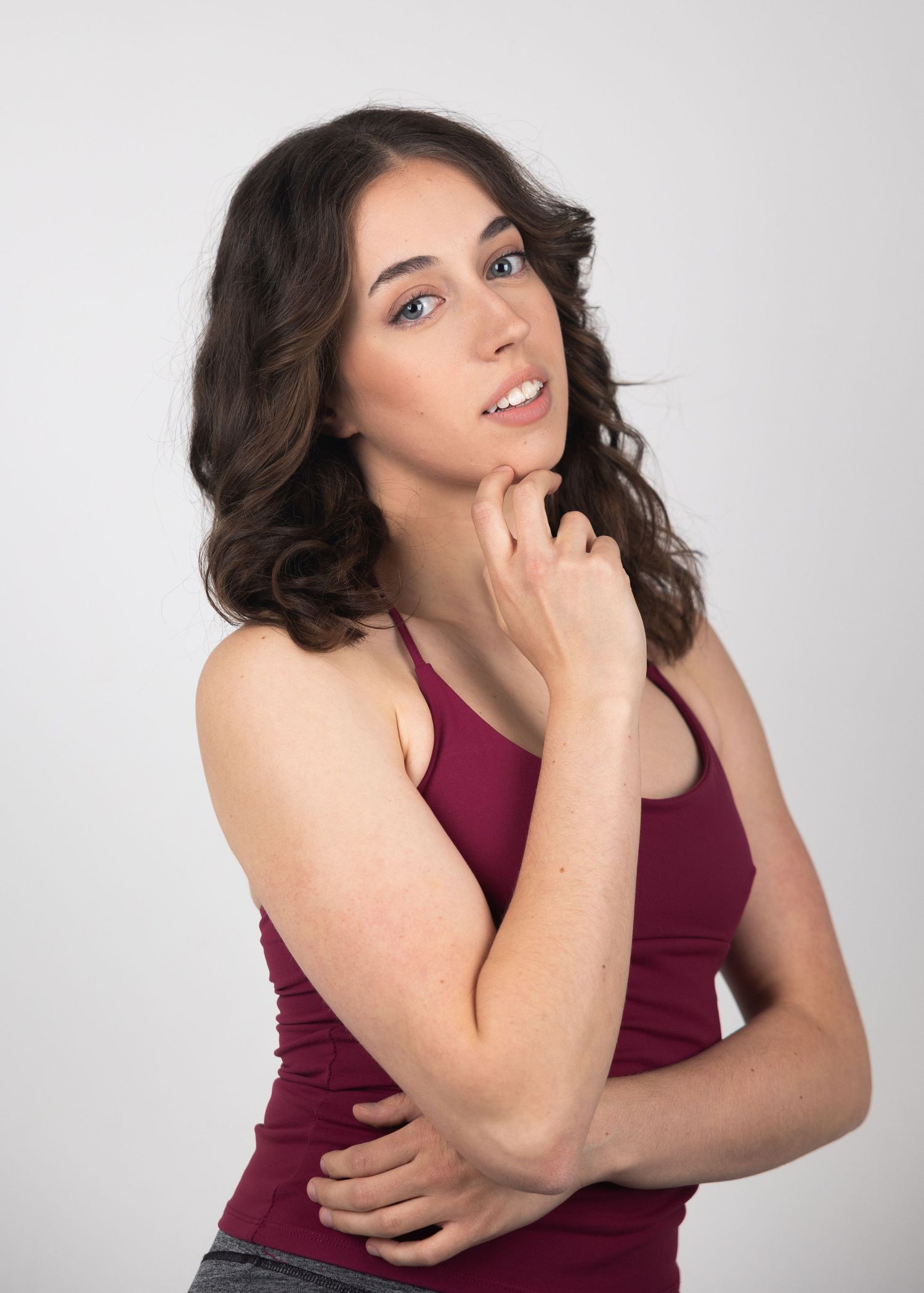Young woman with wavy brown hair in a burgundy tank top posing thoughtfully against a white background.