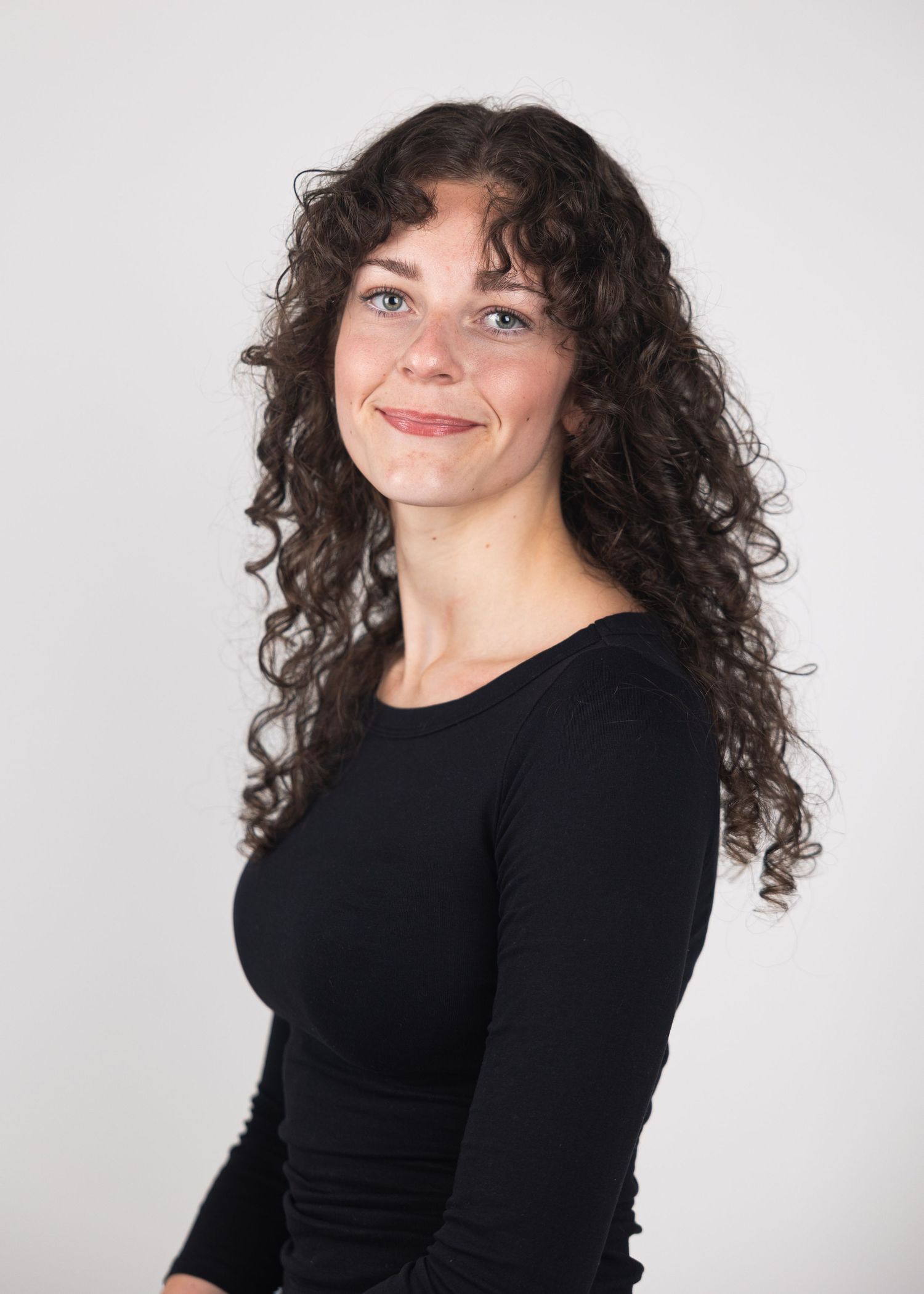 Young woman with long curly dark hair smiling, wearing a black long-sleeve top against a white background.