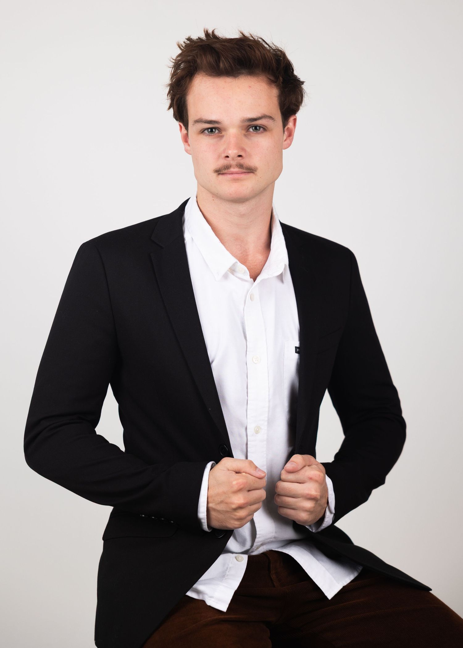 Young man in black blazer and white shirt poses confidently against a light gray background.