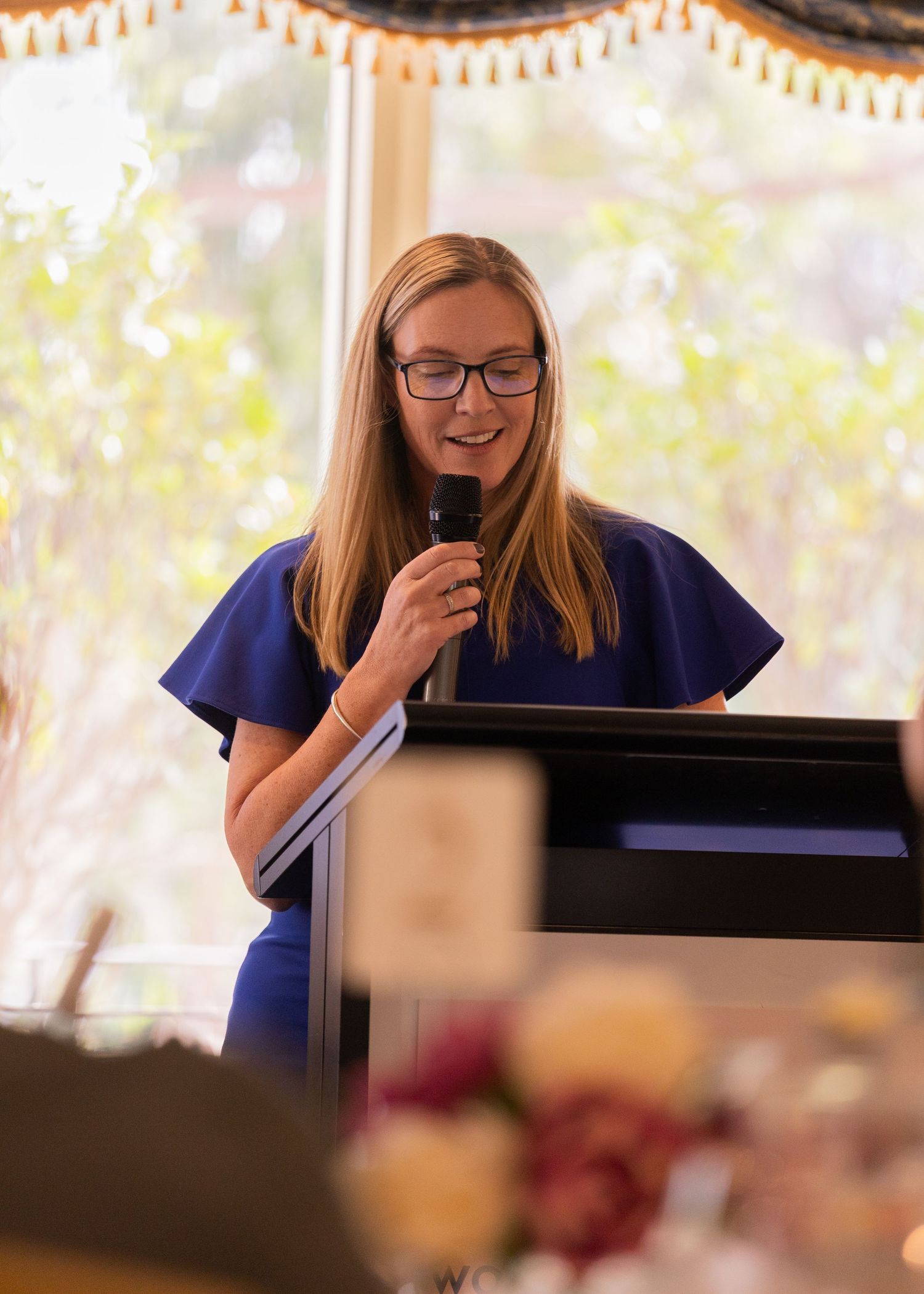 Young woman in blue dress speaking at podium with microphone at formal event.