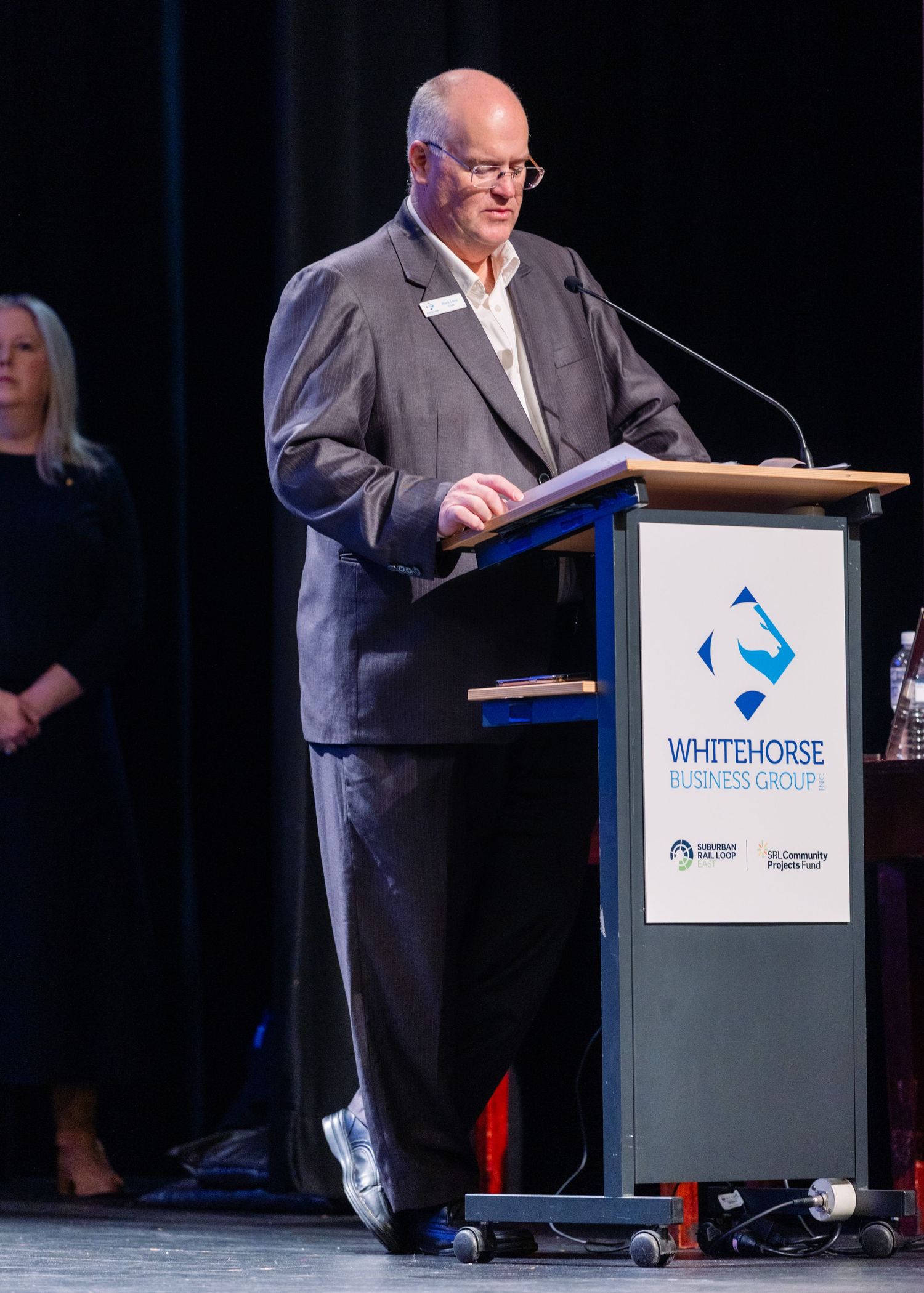 A man in a grey suit speaks at a Whitehorse Business Group podium on stage.