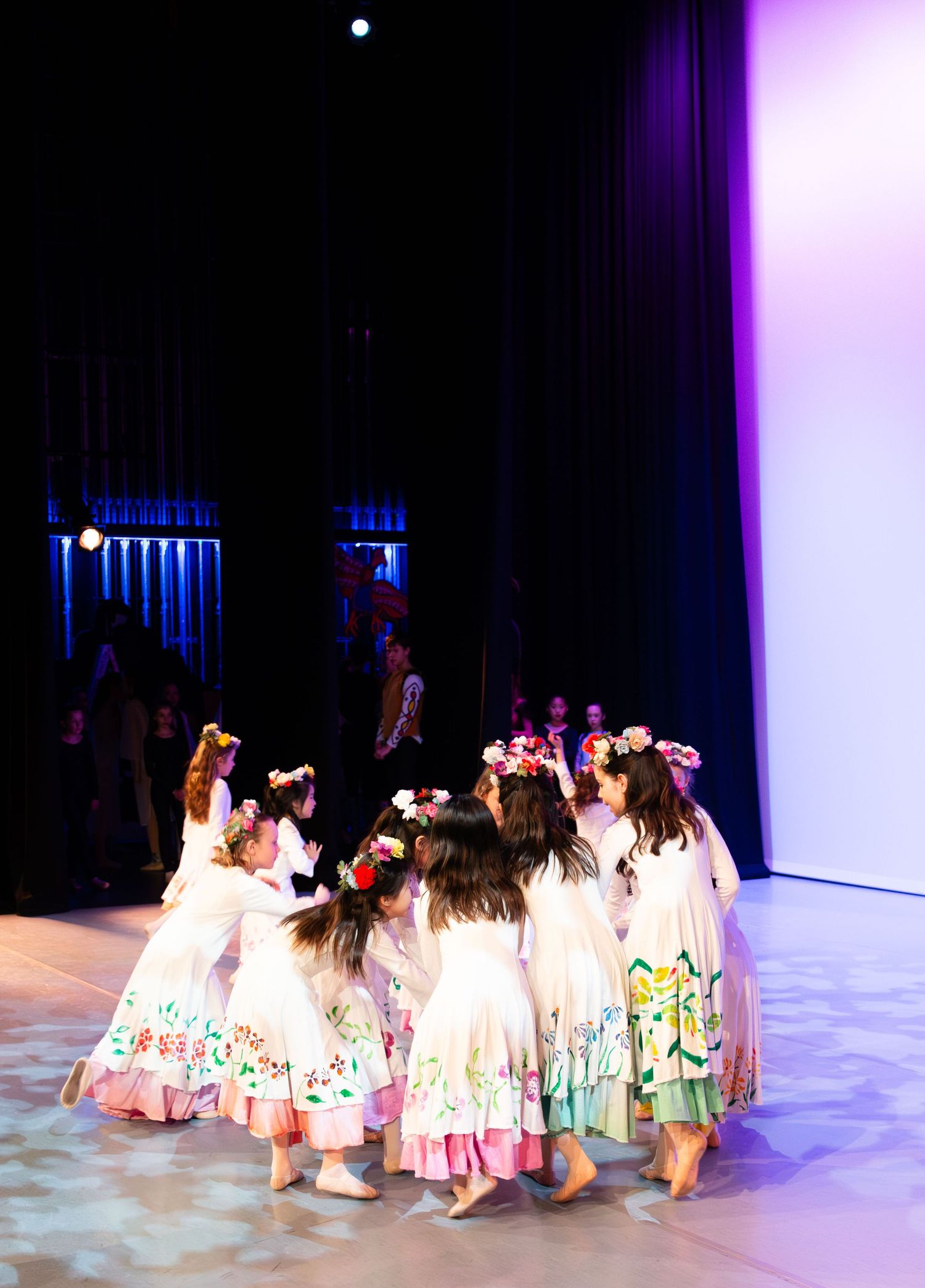 Young girls in floral dresses and flower crowns perform a circle dance on a stage during a recital.