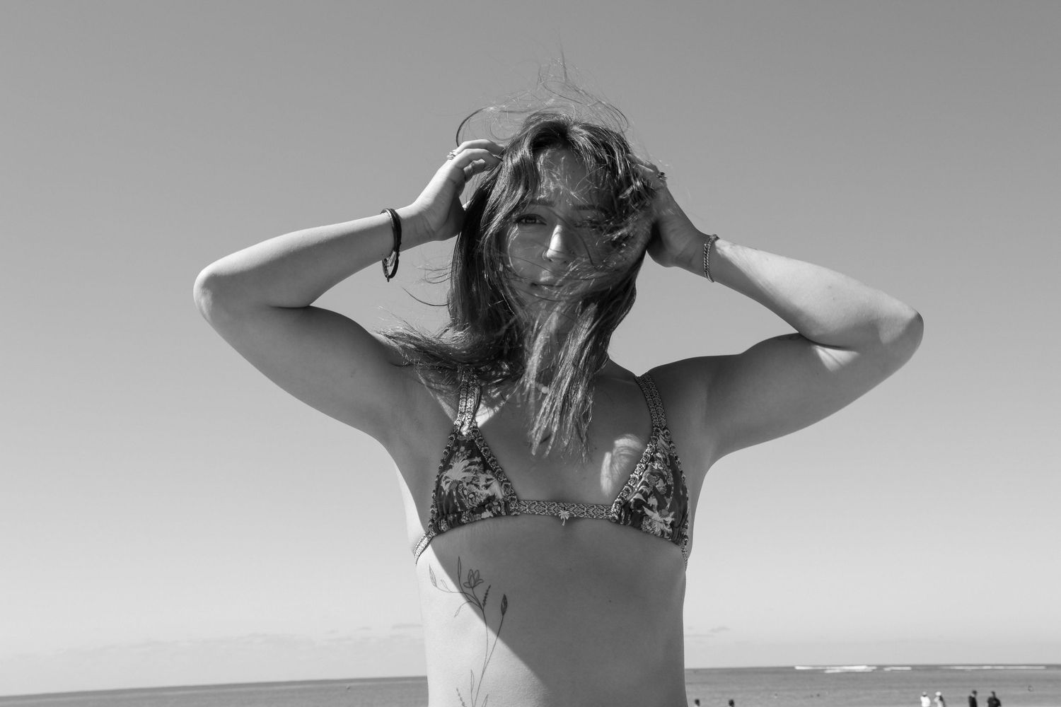Black and white photo of a woman in a bikini at the beach, hands in her hair, with ocean in background.
