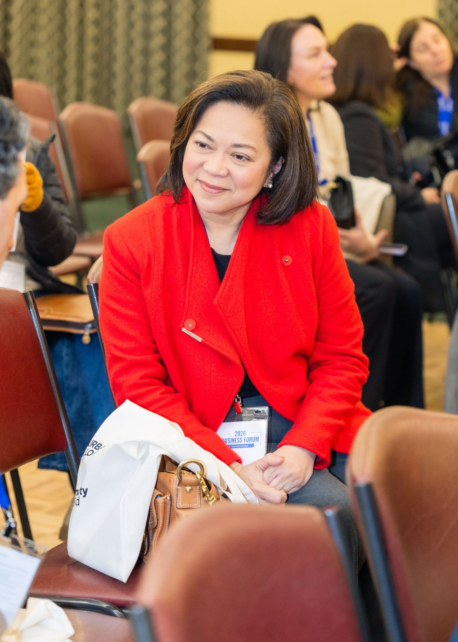 Woman in red blazer smiling at a conference seated in a row of chairs with a name badge.