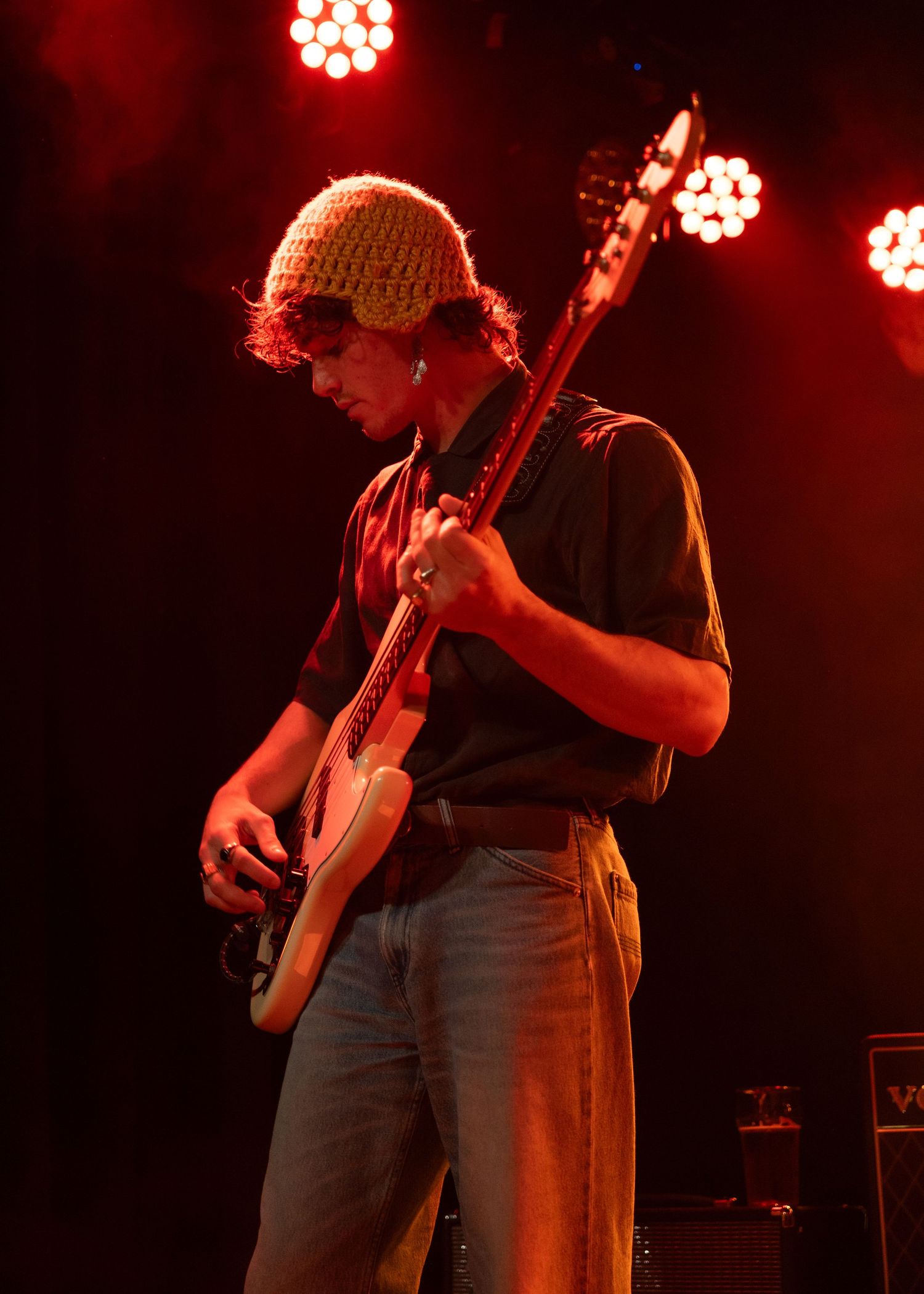 Male guitarist wearing a knit beanie playing electric guitar on stage under red lights.