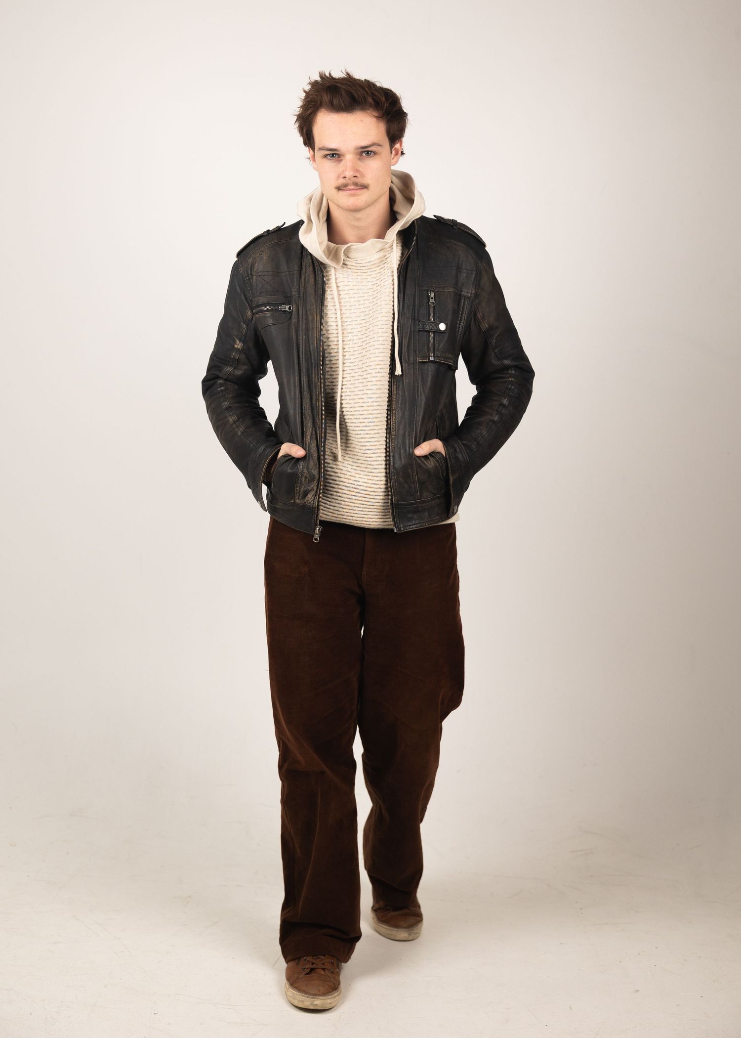 Young man in black leather jacket, cream hoodie, and brown corduroy pants walking forward.