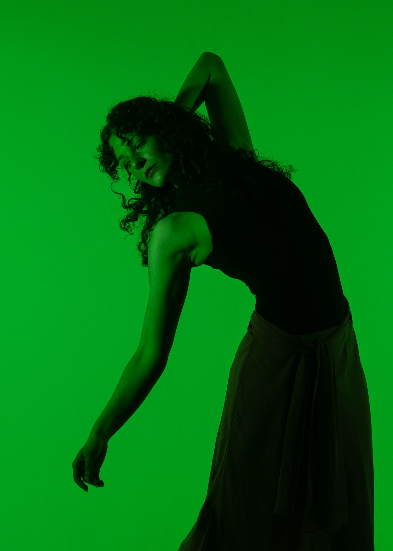 A dancer poses dramatically against a vibrant green background, bending sideways with one arm raised overhead.