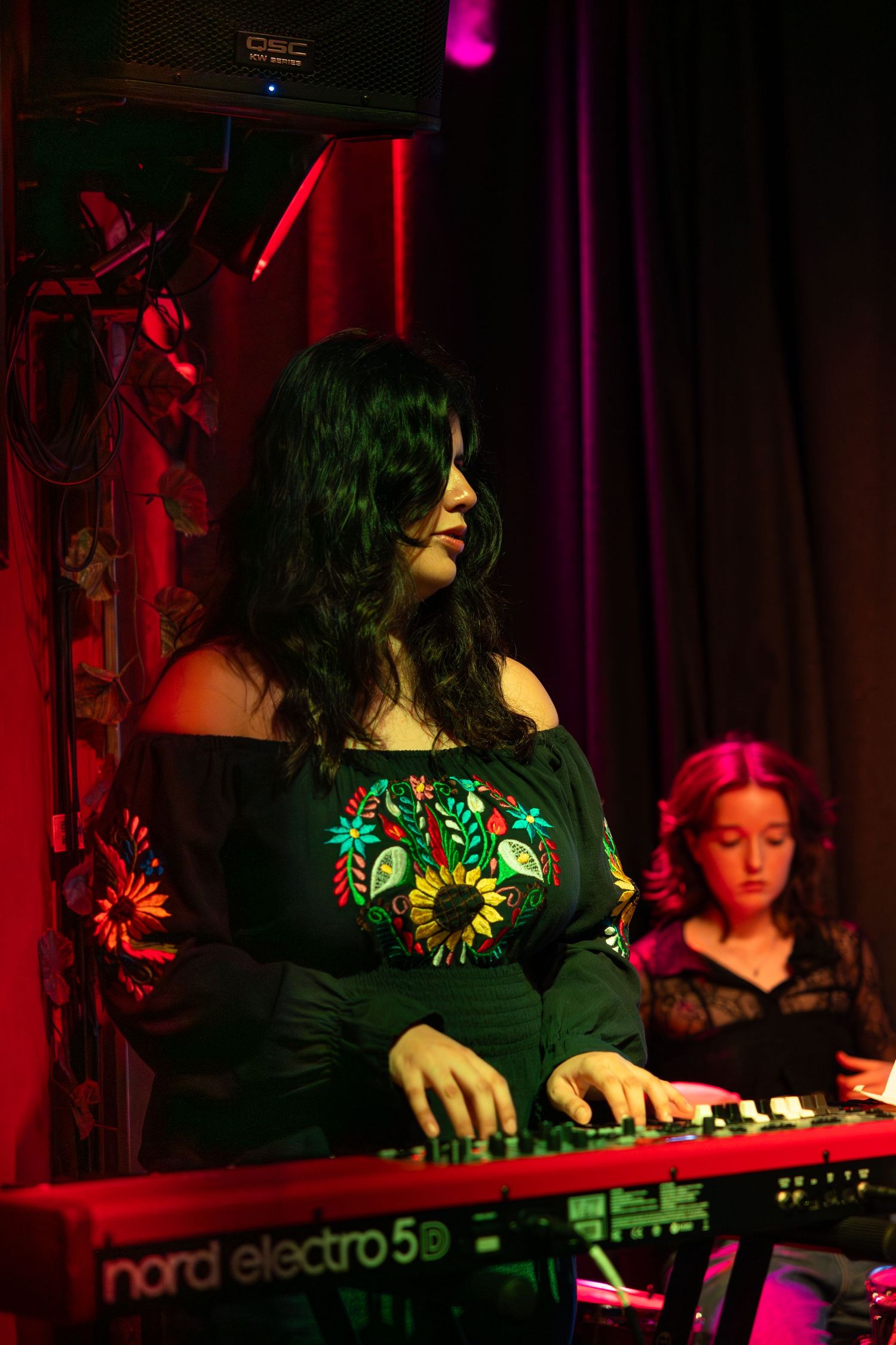 Musician performs on keyboard instruments under dramatic red and purple stage lighting at a live music venue.