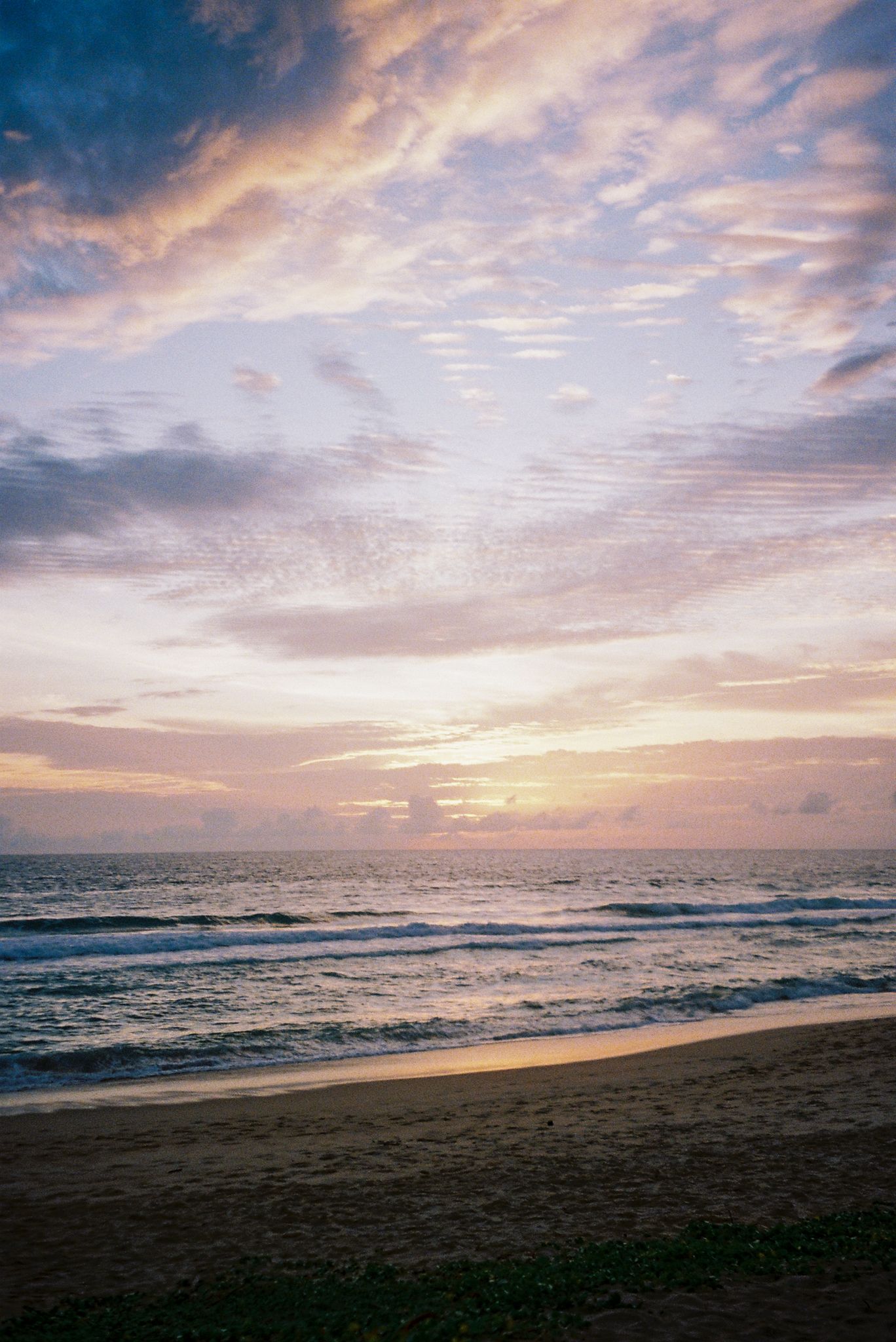 A panoramic sunset view over the ocean with dramatic clouds and waves rolling onto a dark sandy beach.
