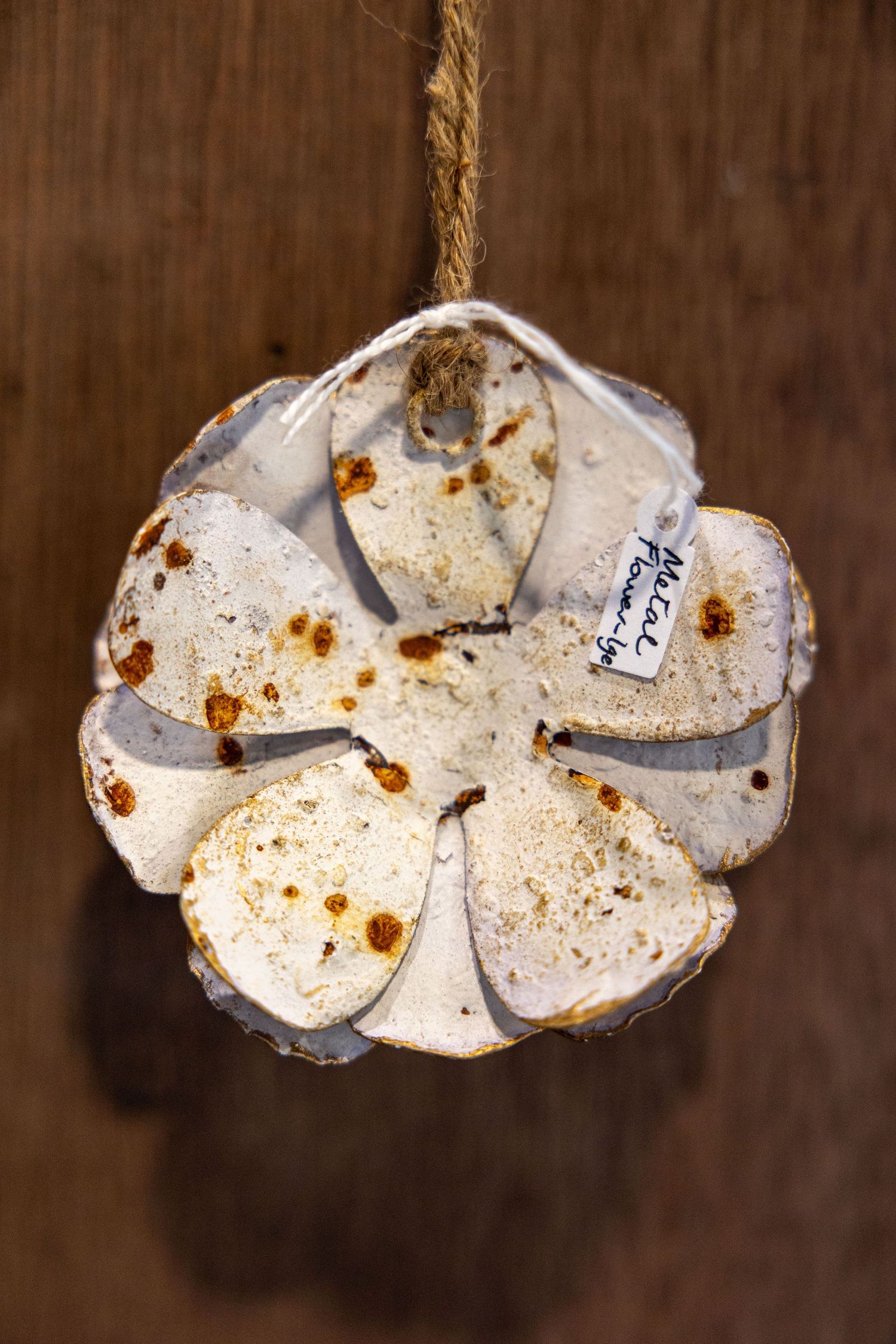 Rustic white metal flower ornament with rust spots, hanging by twine rope on wooden background.