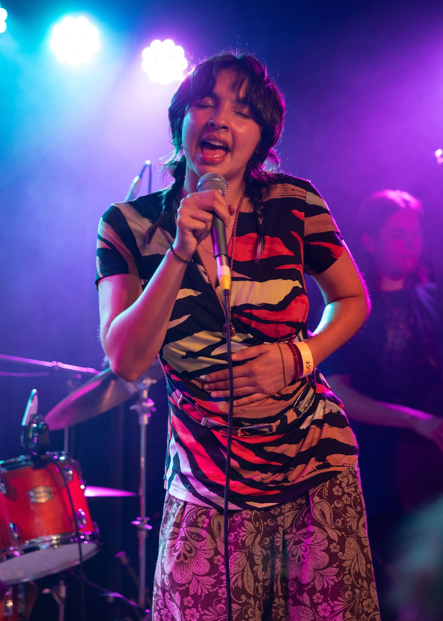 Woman in colorful zebra-print top singing into microphone on stage with purple and blue stage lighting.