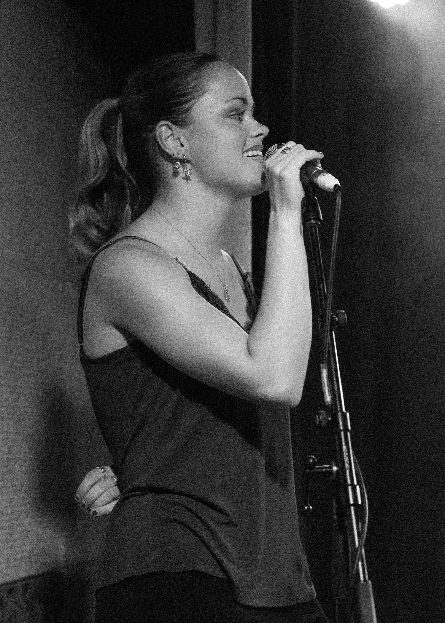 Black and white photo of a woman singing into a microphone on stage with a ponytail and sleeveless top.