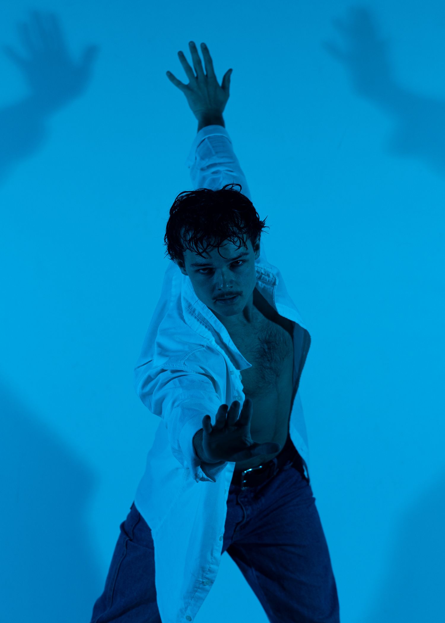 Person in white open shirt and jeans poses dramatically against blue background with shadow hands.