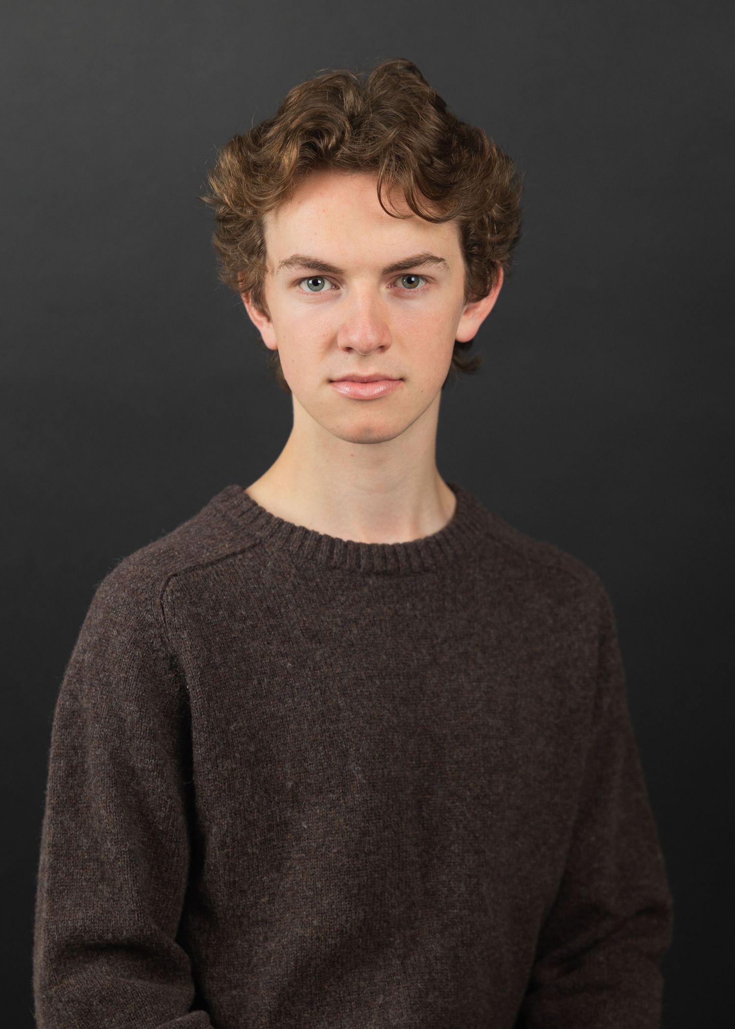 Young man with curly brown hair wearing a dark brown knit sweater poses for a professional headshot photo.