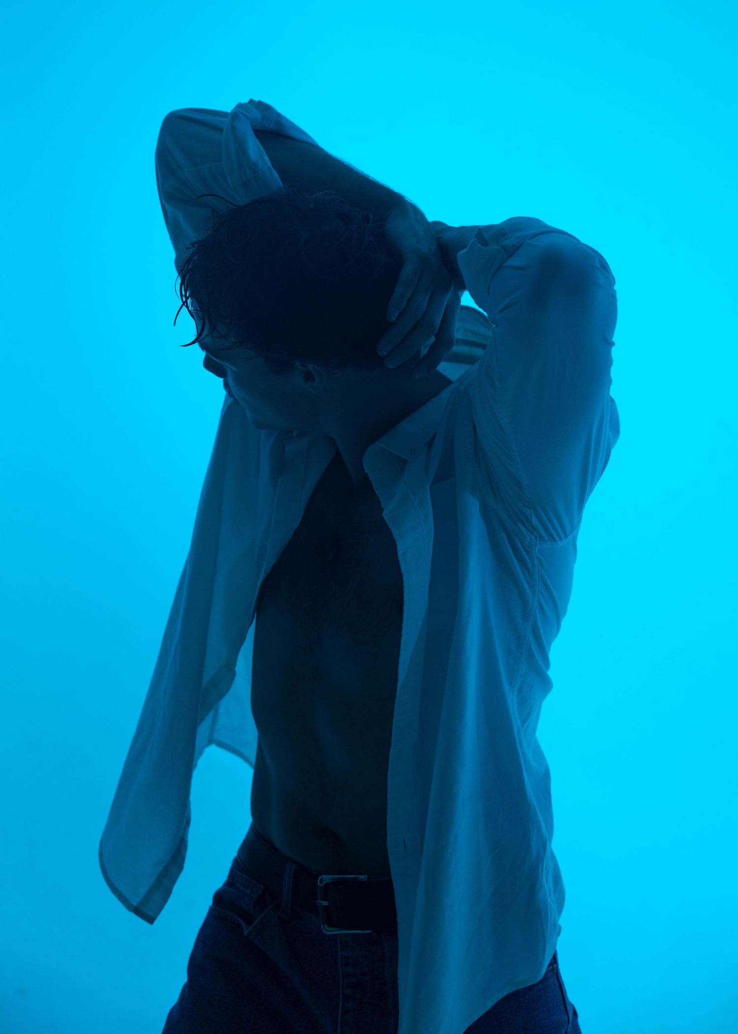 Male dancer poses dramatically with hands behind head in an open white shirt under blue studio lighting.
