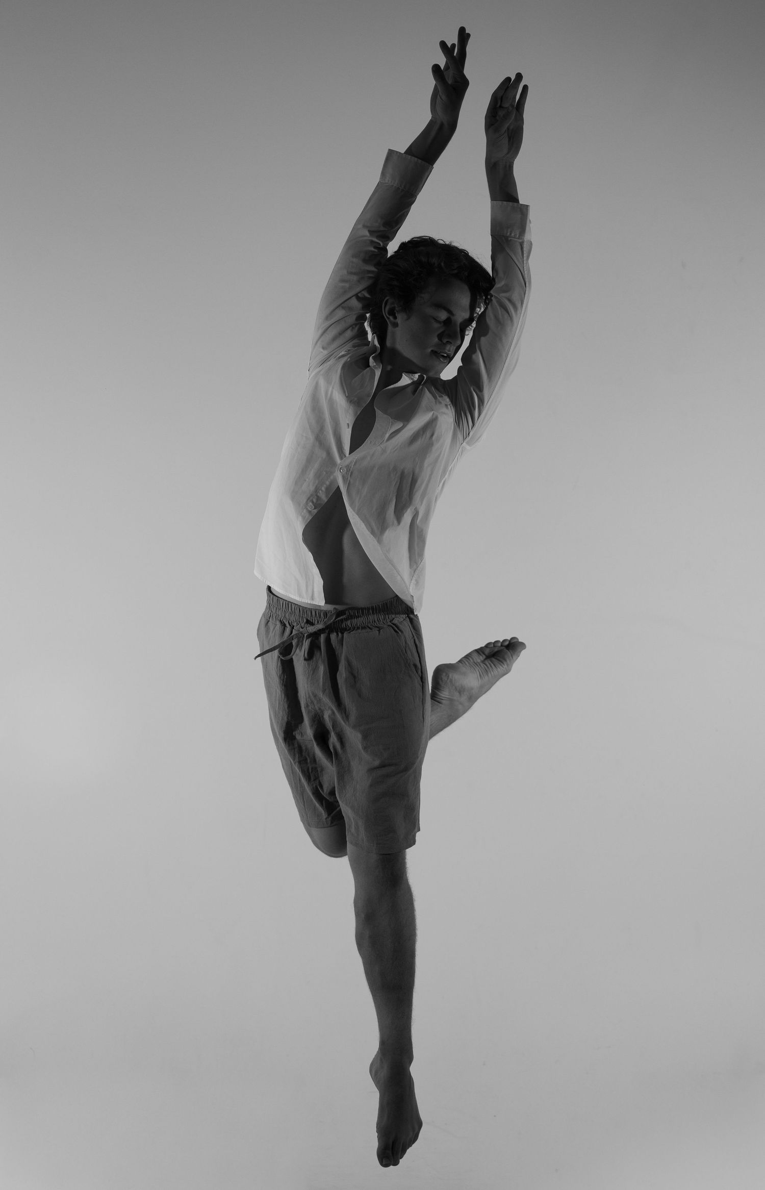 Black and white photo of a dancer mid-leap, arms raised overhead, one leg bent, wearing shorts and a light top.