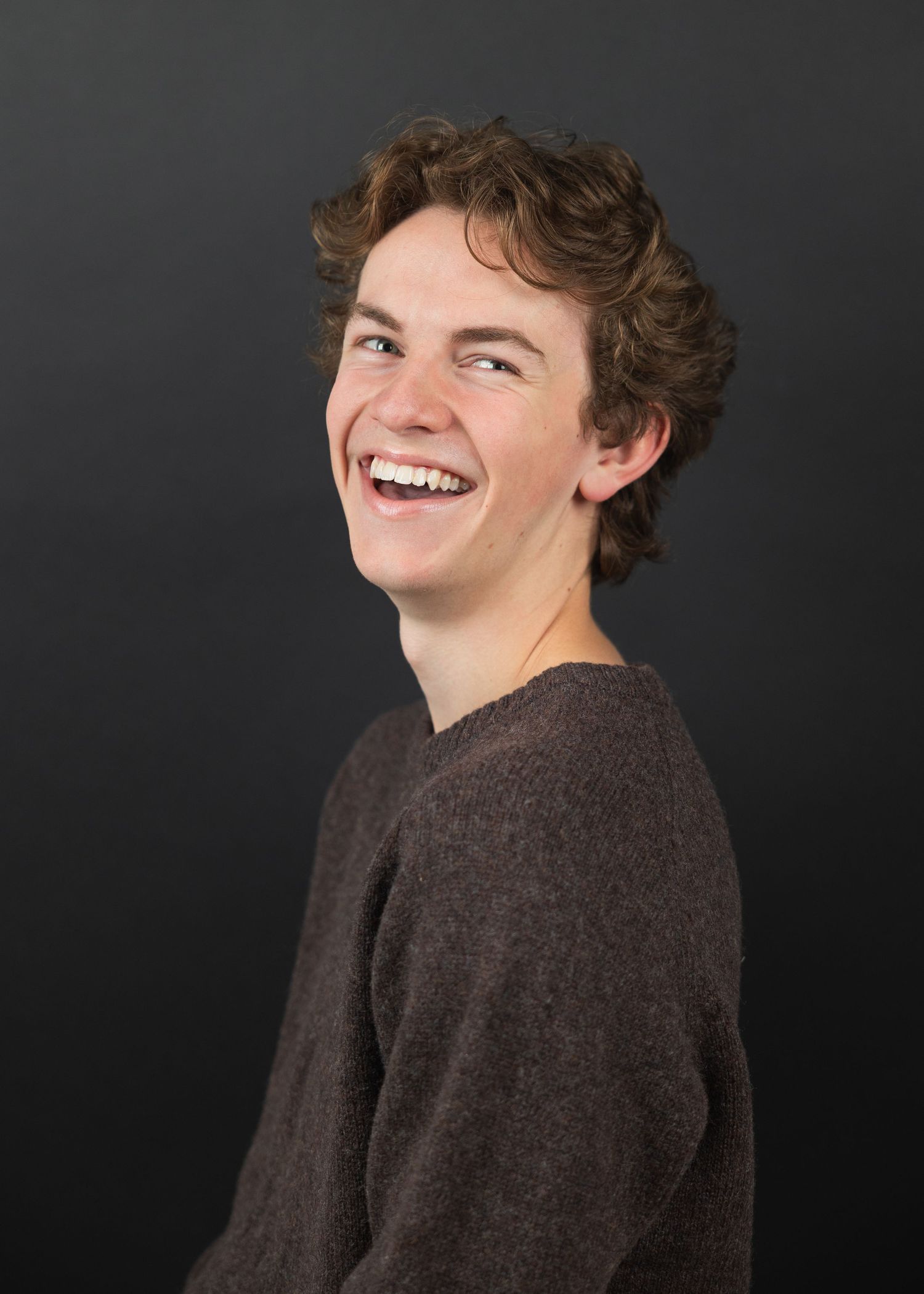 A young person with curly hair laughs joyfully, wearing a brown sweater against a dark gray background.