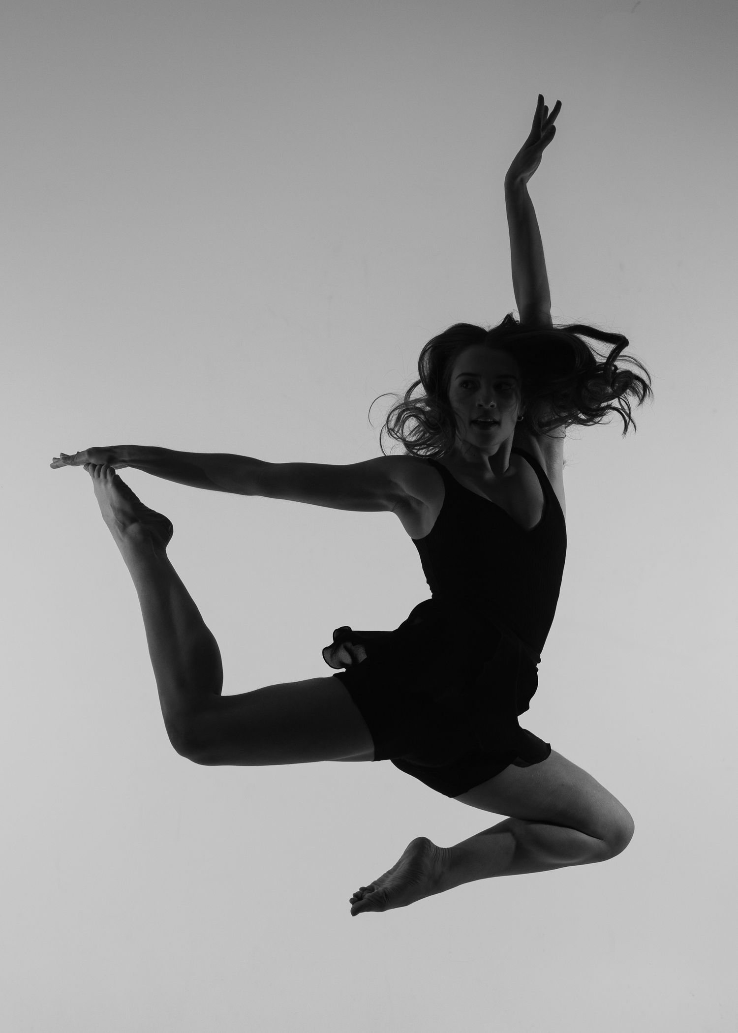 Black and white photo of a female dancer mid-air, gracefully holding her leg in a stunning jump pose.