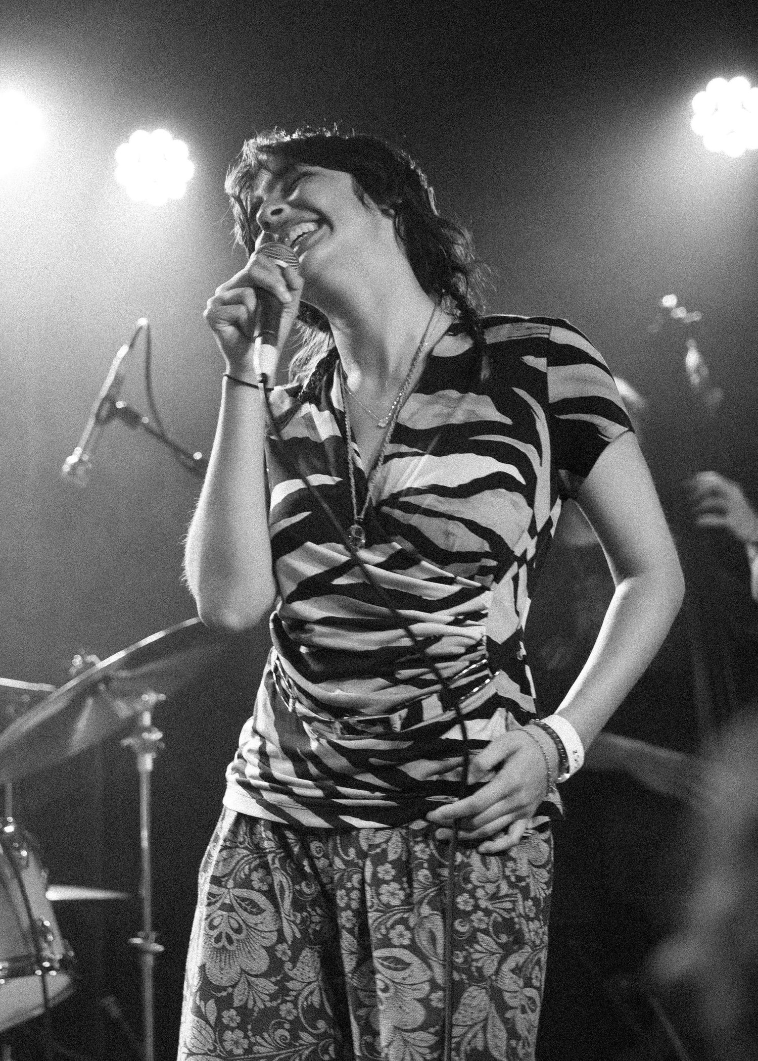 Black and white photo of a woman singing into a microphone on stage, wearing a striped top and floral skirt.