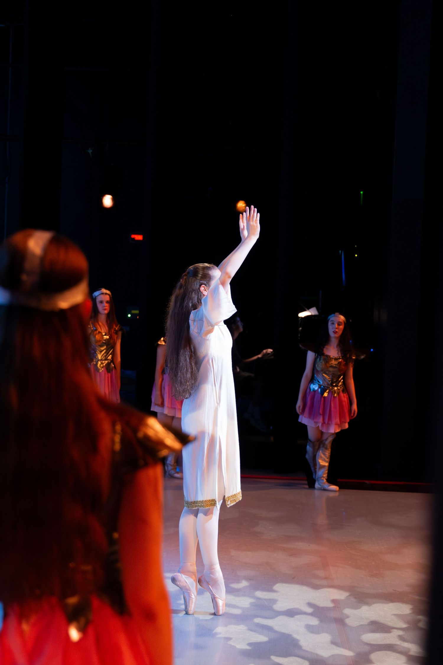 Ballet dancers perform on stage, with a soloist in white dress en pointe raising her arm gracefully under dramatic lighting.