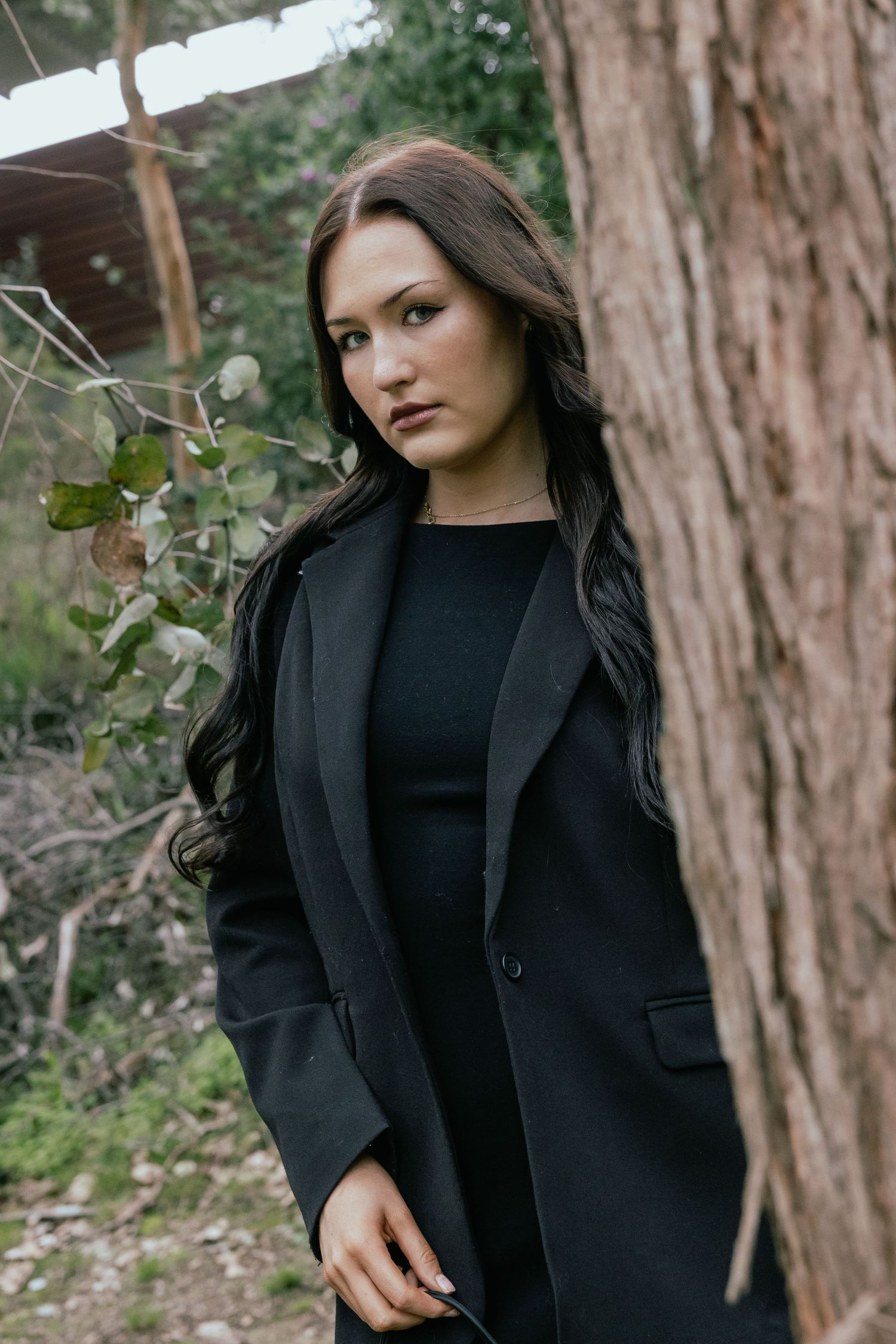 A young woman with long dark hair wearing a black coat stands near a tree in a wooded outdoor setting.