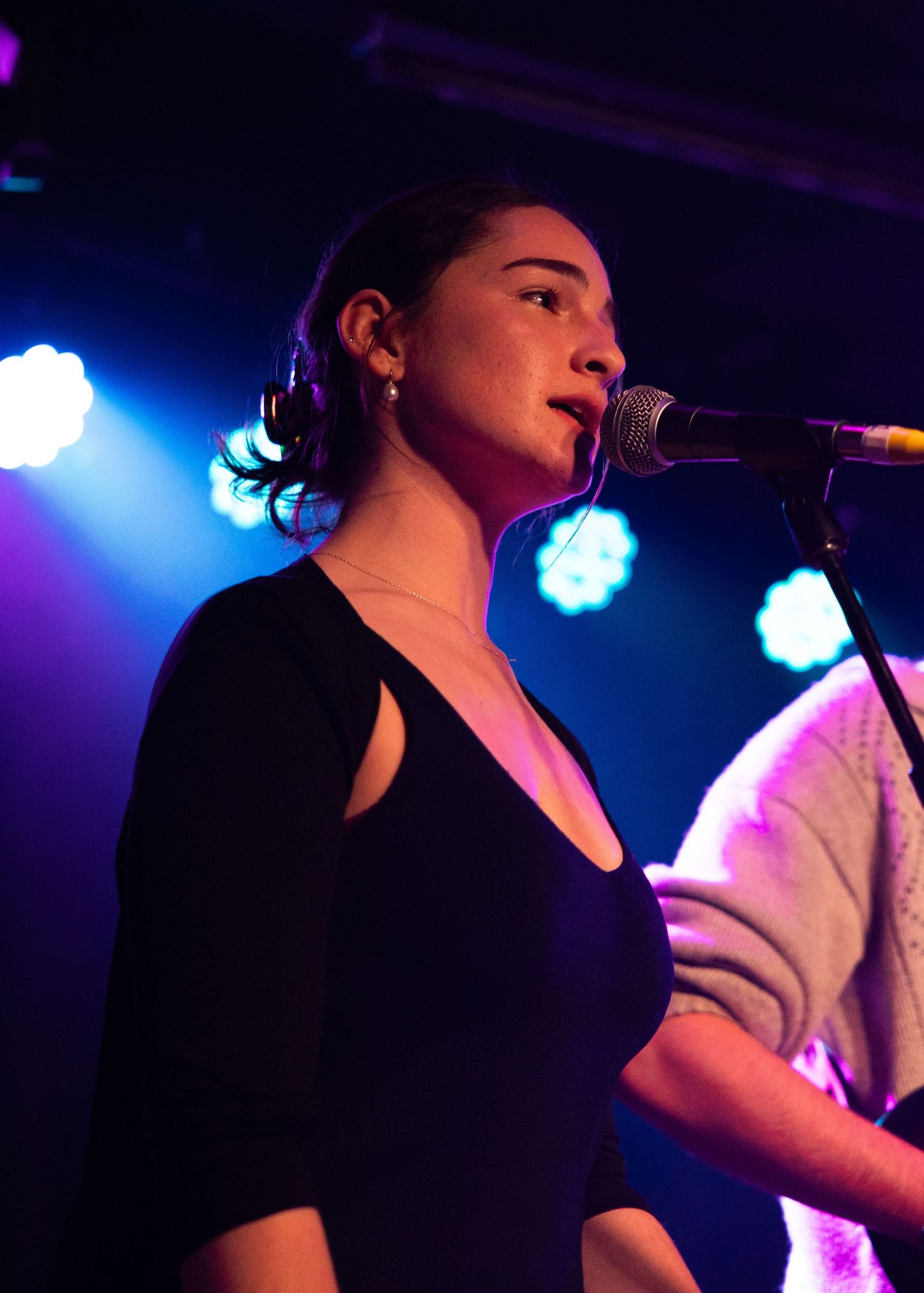 Young woman in black dress singing into microphone on stage with blue and purple stage lights behind her.