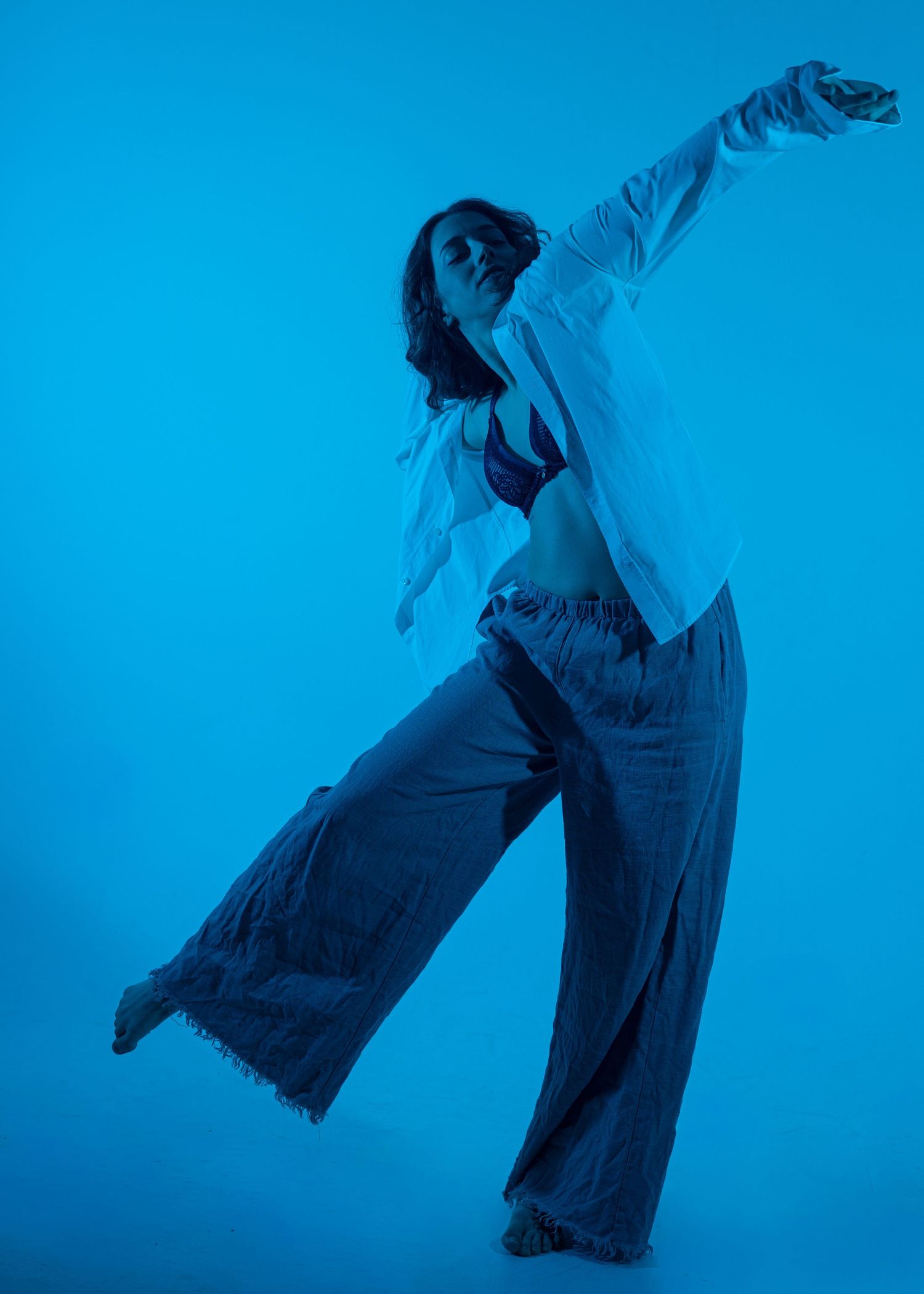 A female dancer poses gracefully on a blue background, wearing wide-leg pants and an open white shirt, arm extended.