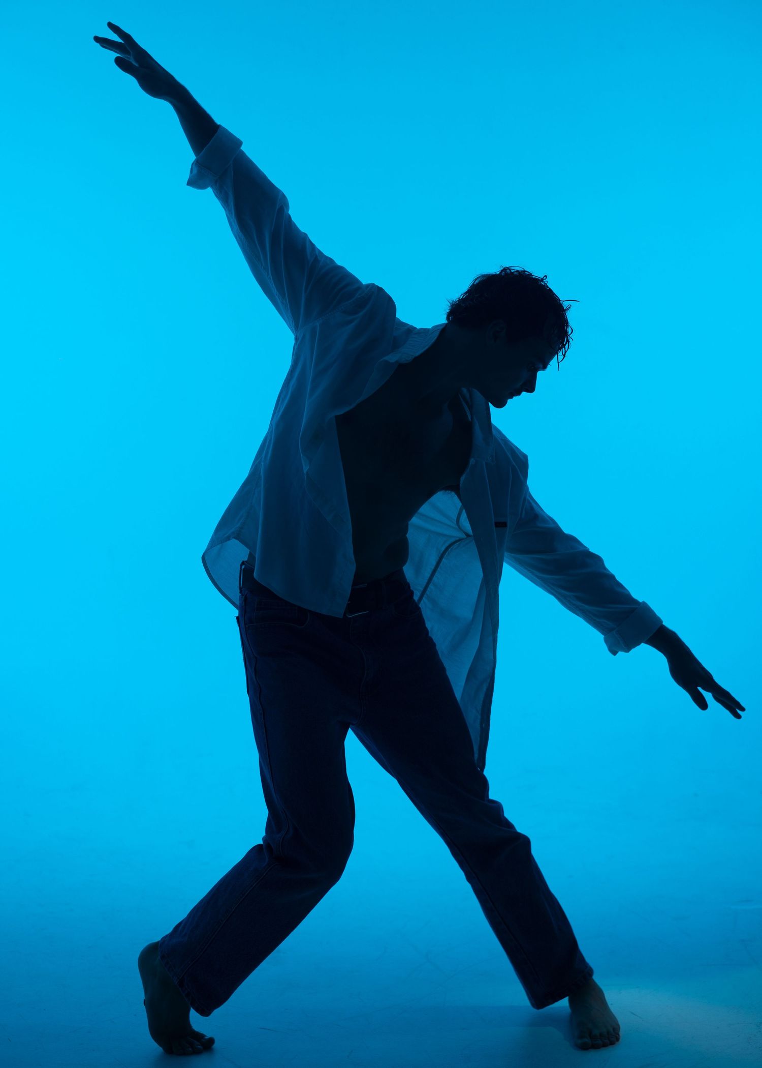 Male dancer performs dramatic contemporary pose in white shirt under blue studio lighting.