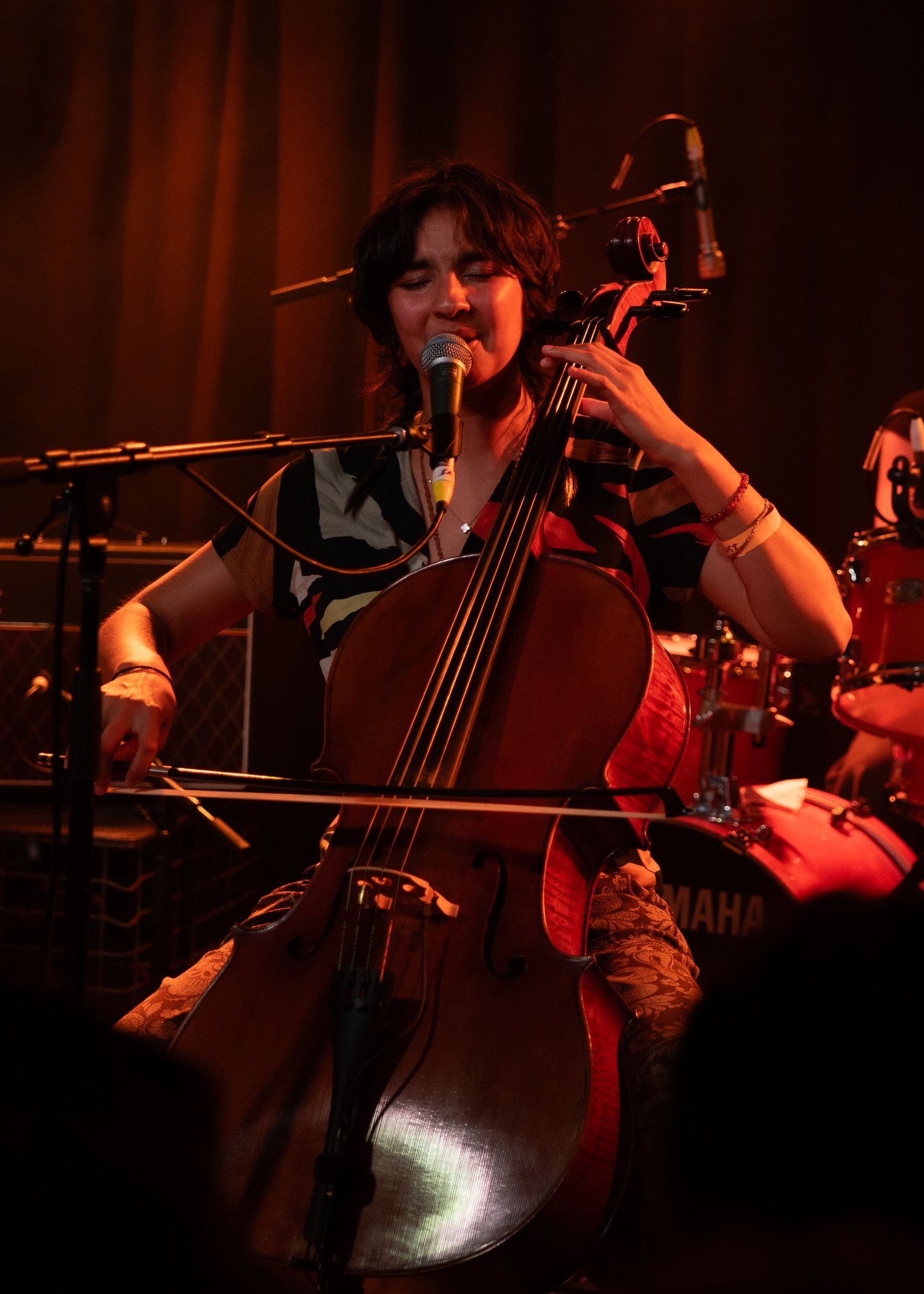 Female cellist performing on stage, playing cello and singing into microphone under warm red stage lighting.