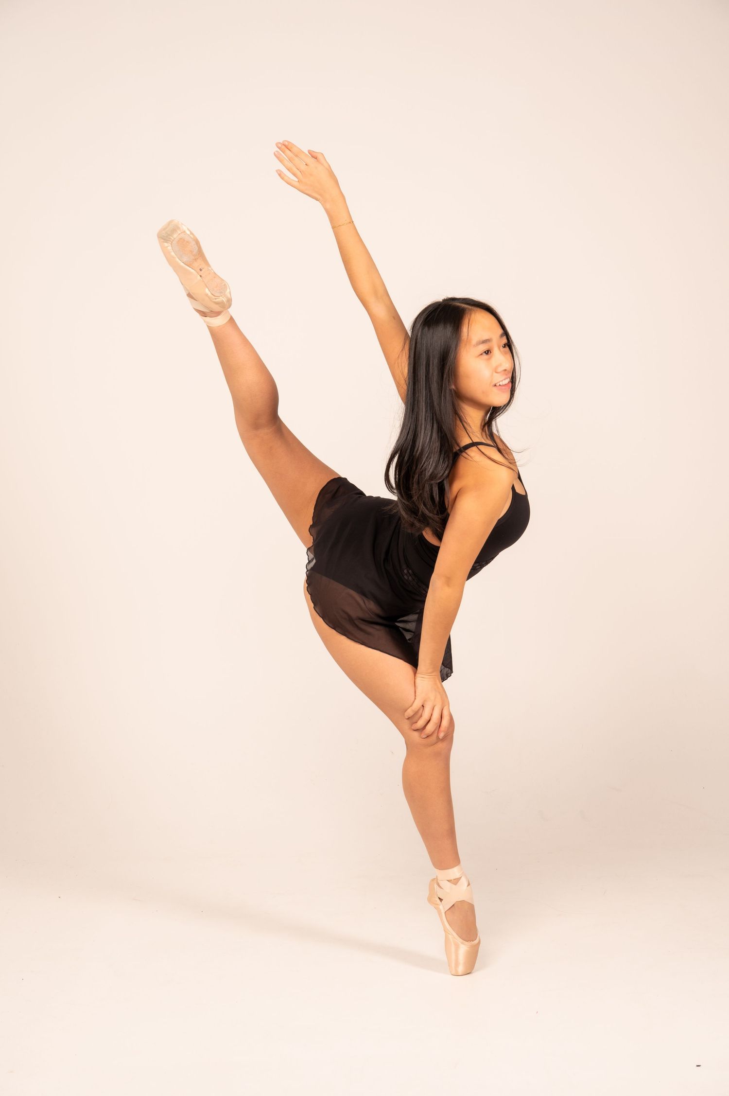 Dancer in black leotard performs high leg extension against white backdrop in graceful dance sequence.