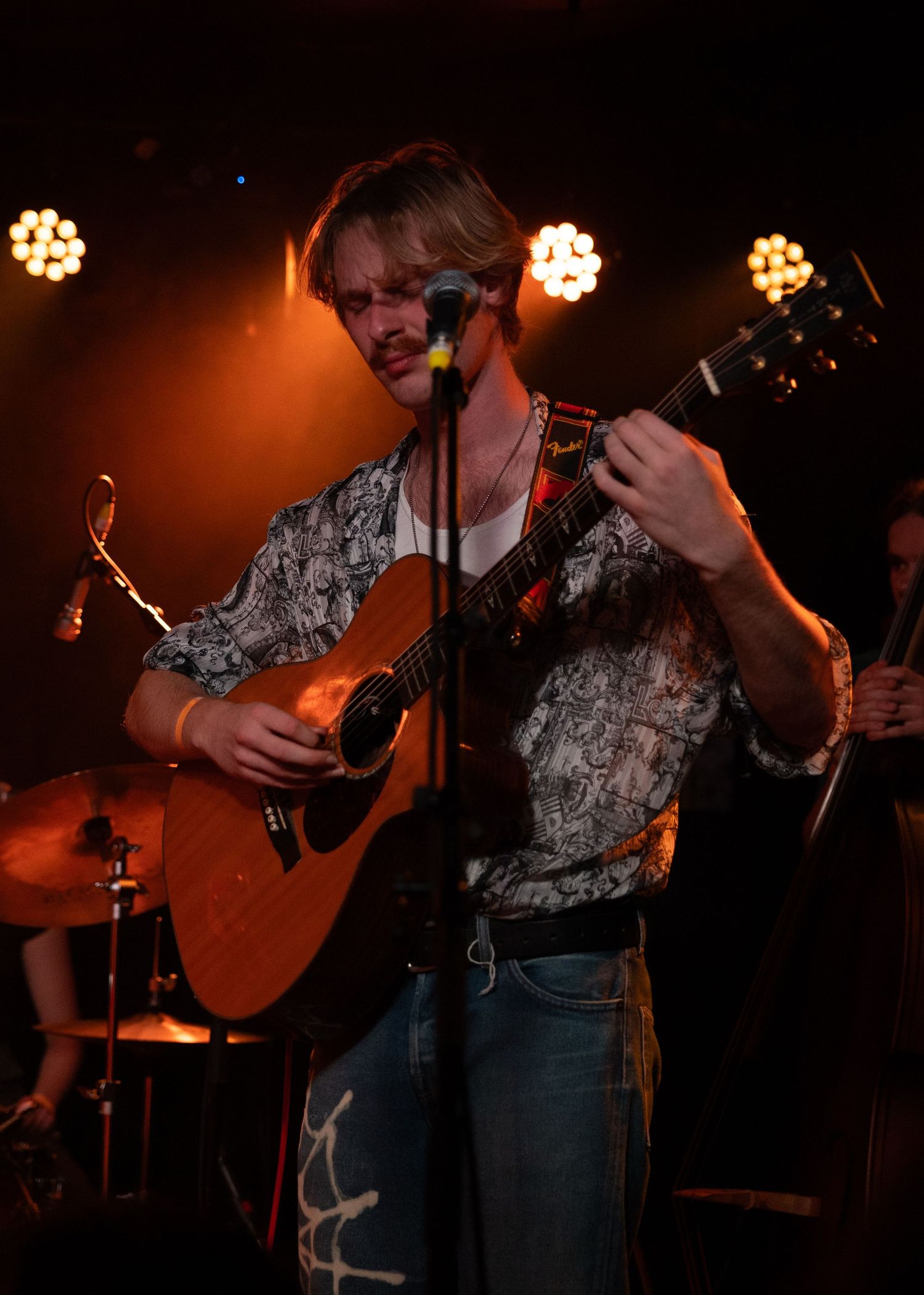 Young male musician playing acoustic guitar on stage under warm orange lights during a live performance.