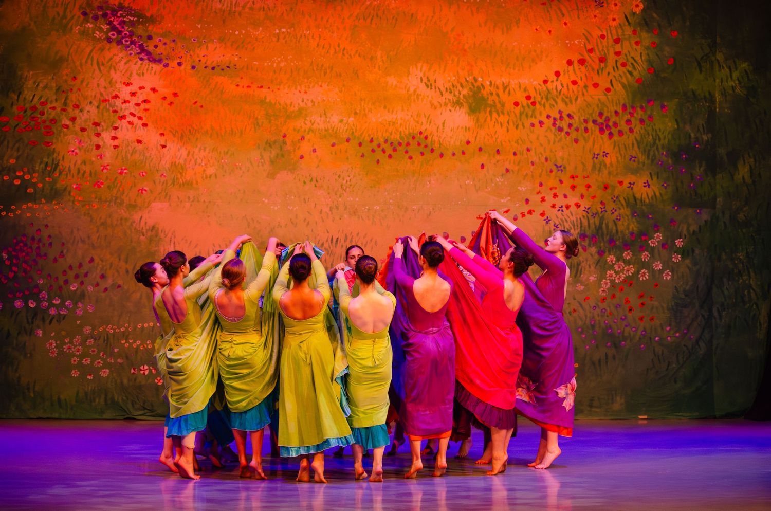 Dancers in vibrant yellow and purple dresses perform in formation against an orange backdrop on stage.