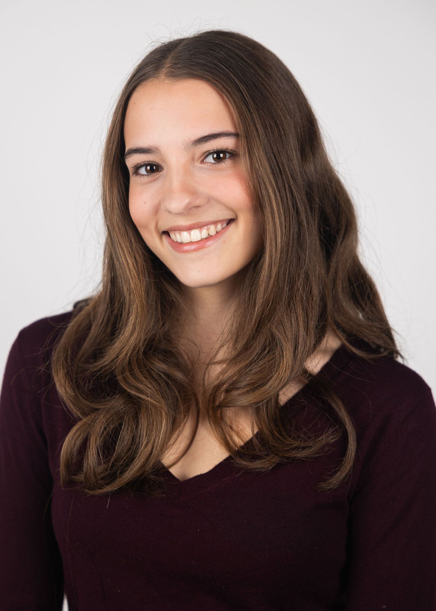 Smiling young woman with long brown wavy hair in a burgundy v-neck top poses for a professional headshot.