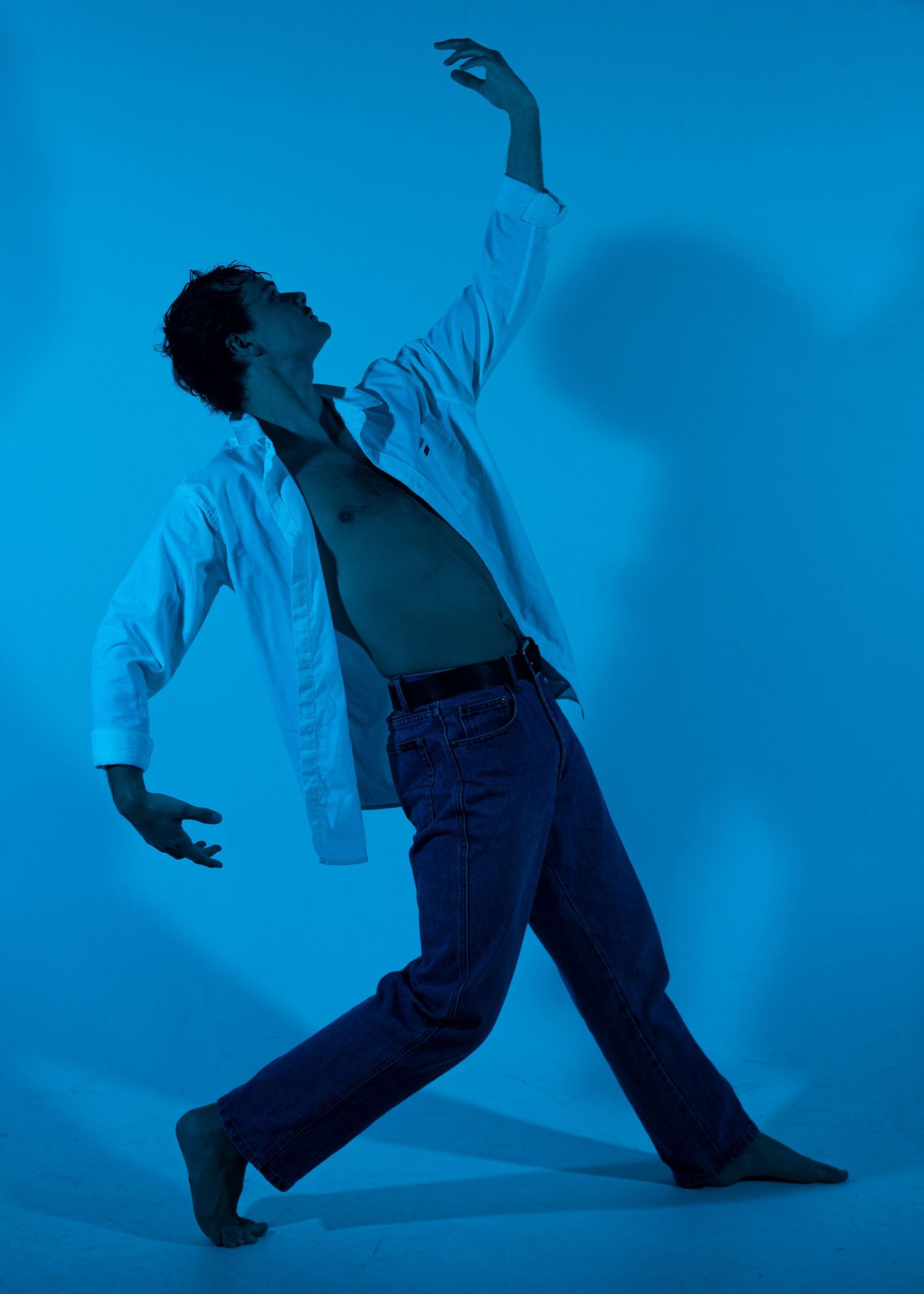 Male dancer in open white shirt and jeans striking dramatic pose with raised arm against blue background.